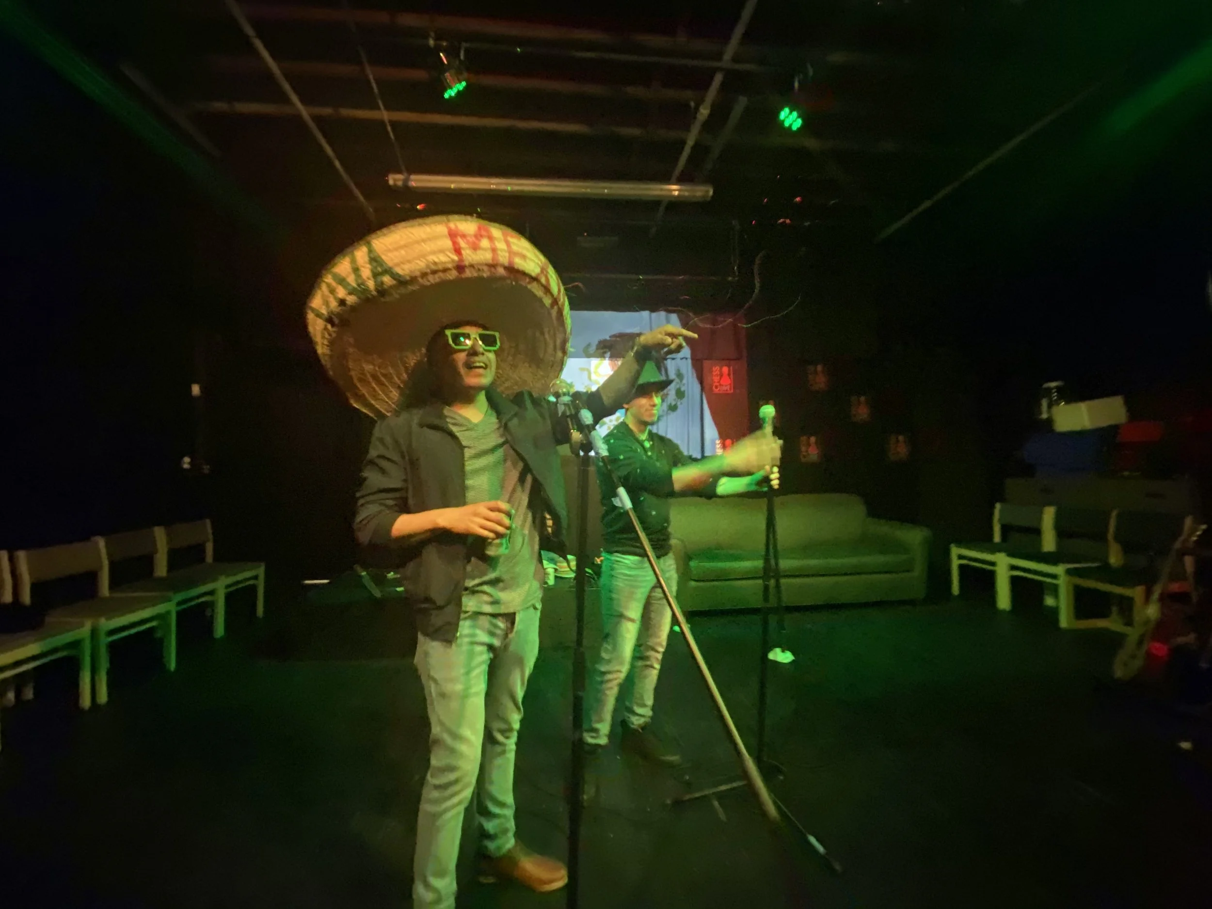 Two men performing on stage with microphones, one wearing green sunglasses and a large sombrero, the other pointing. The background has a screen and black walls with chairs along the sides.