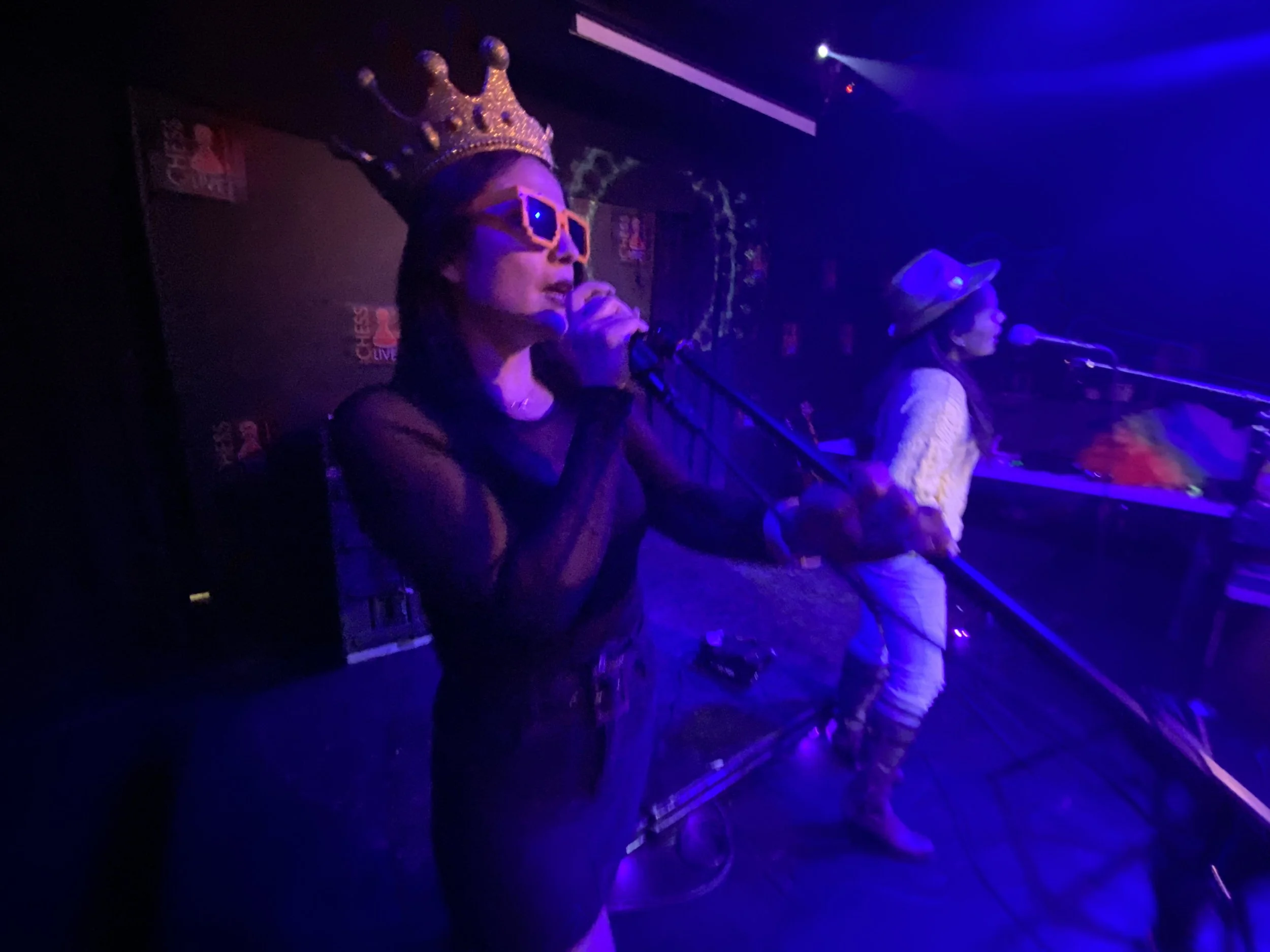 Woman wearing a crown and sunglasses singing into a microphone on stage with another woman playing keyboard, under purple lighting at a live music venue.