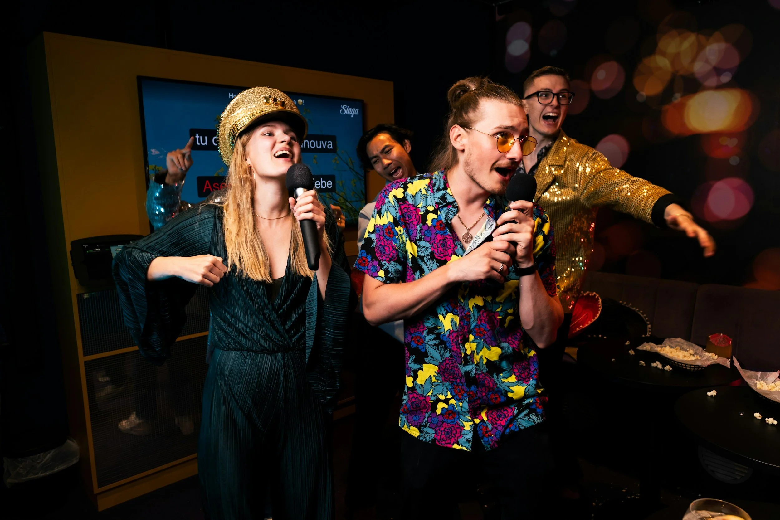 Group of young adults singing and dancing at a karaoke party, holding microphones, with party decorations and snacks on the table.