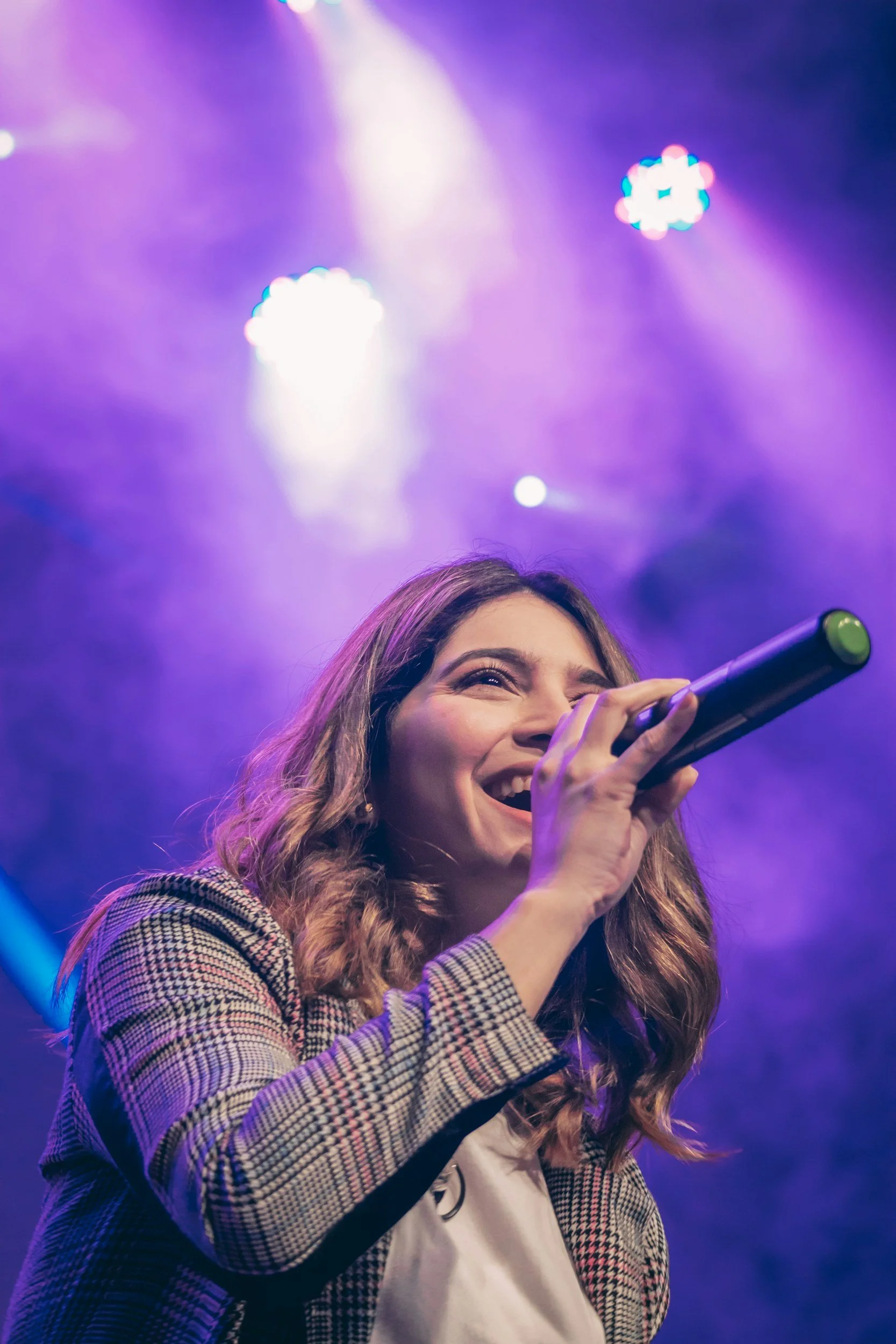 A woman with wavy hair holding a microphone and smiling on stage with purple and blue lighting