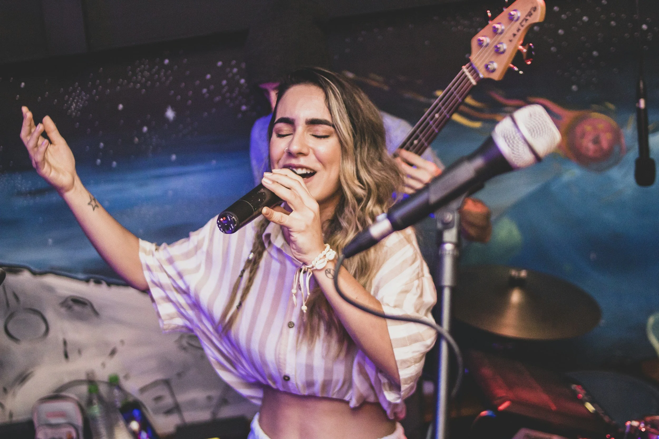 A young woman singing passionately into a microphone with her eyes closed and hand raised, while a musician in the background plays a bass guitar, at a lively music venue with colorful and space-themed decor.