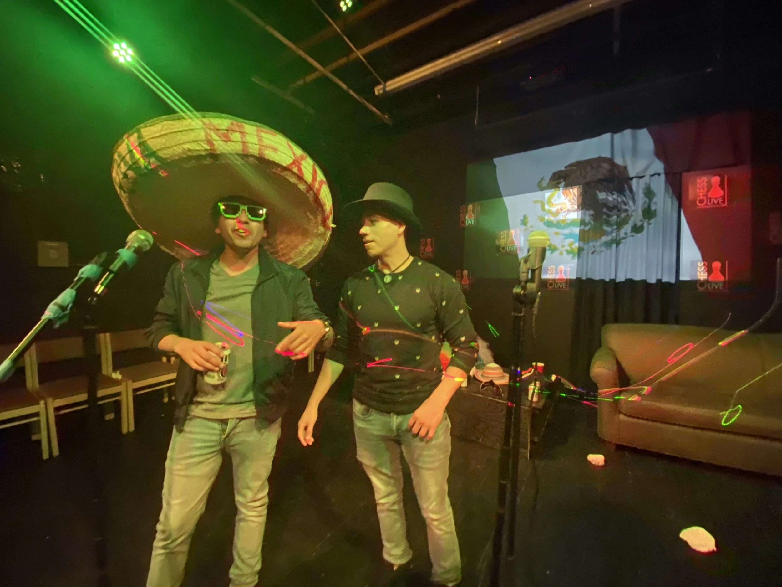 Two men standing on a stage with a Mexican theme, one wearing a large sombrero and sunglasses, the other wearing a hat, surrounded by microphones, with a screen behind displaying Mexican imagery and a logo that reads 'Chess Live'.