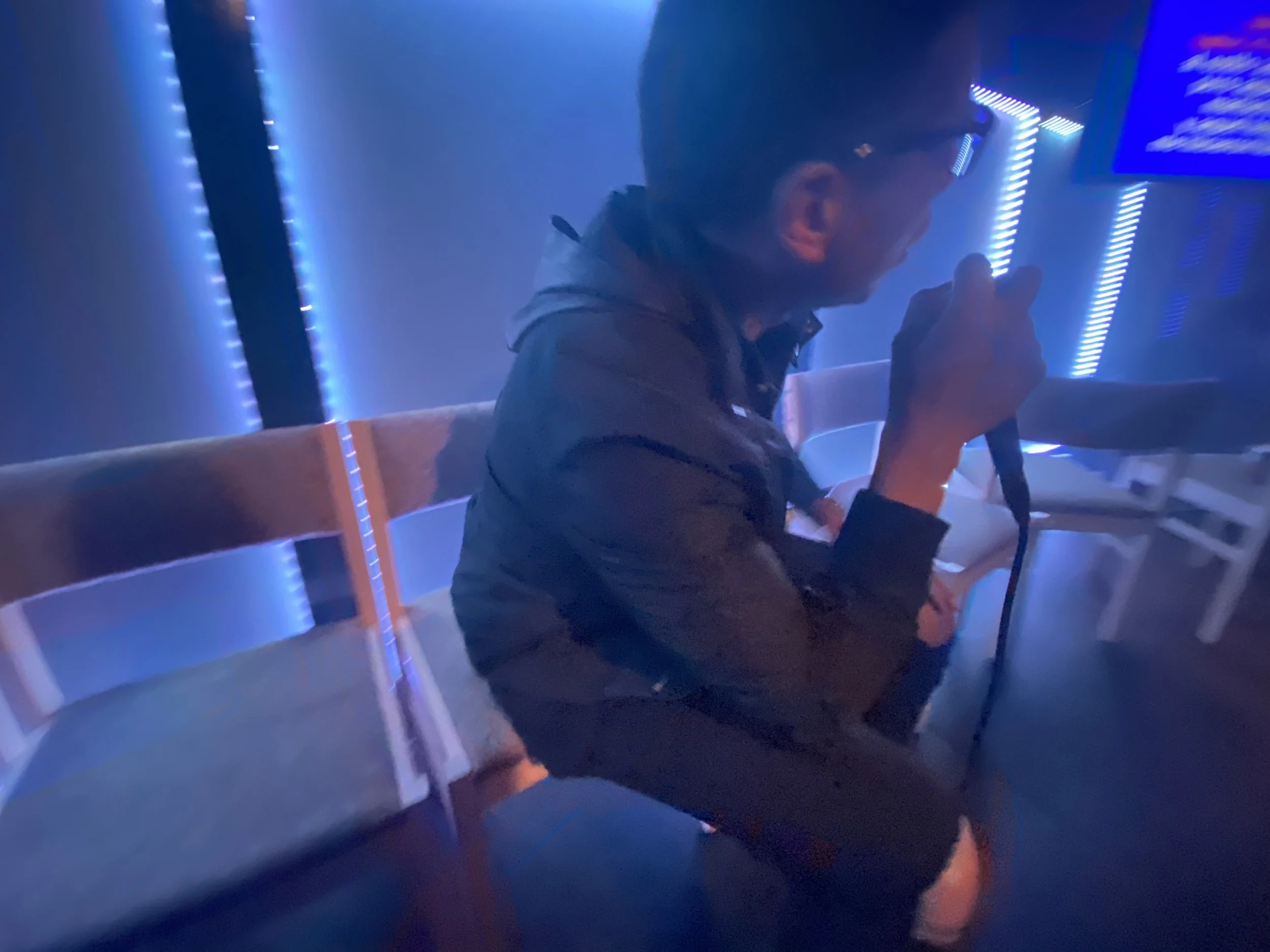 A person sitting on a bench in a dark room with blue neon lights, holding a microphone and singing.