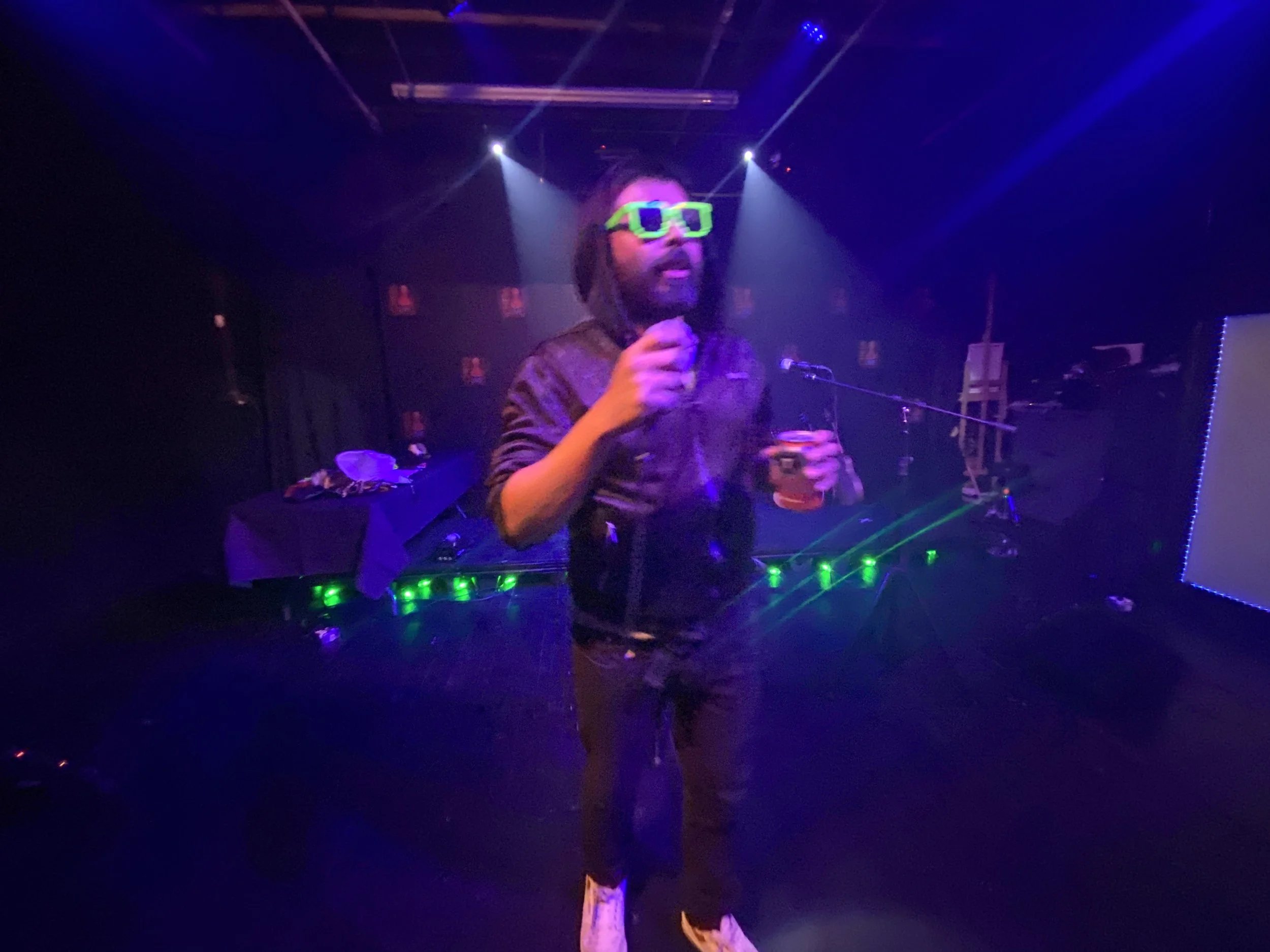 A man with sunglasses holding a microphone and a drink, standing on a stage with colorful lighting and a DJ setup in the background.