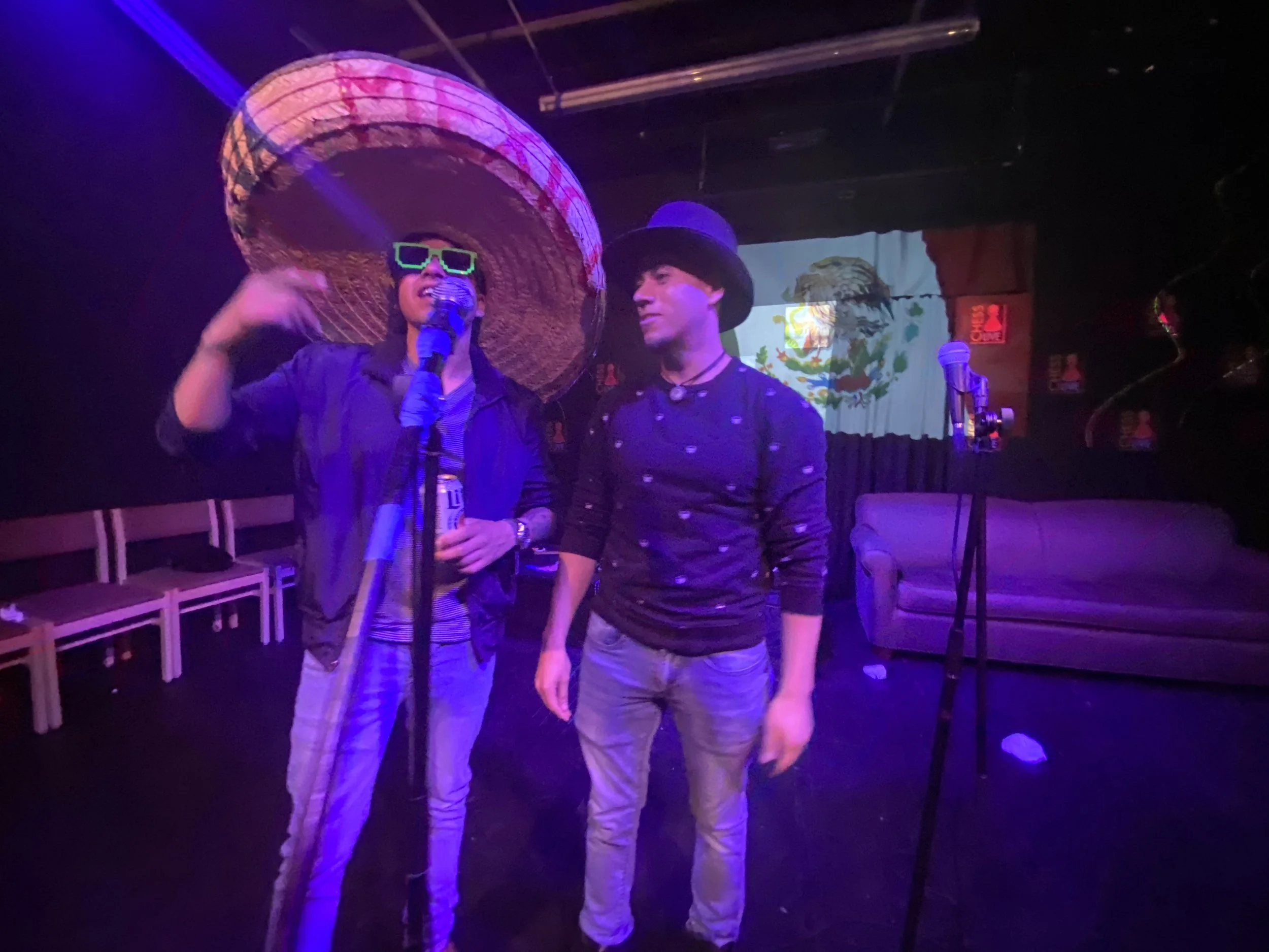 Two young men standing on a stage, one wearing large sombrero and sunglasses, speaking into a microphone, the other wearing a black hat and patterned shirt, standing next to him. Stage lights and Mexican flags in the background.