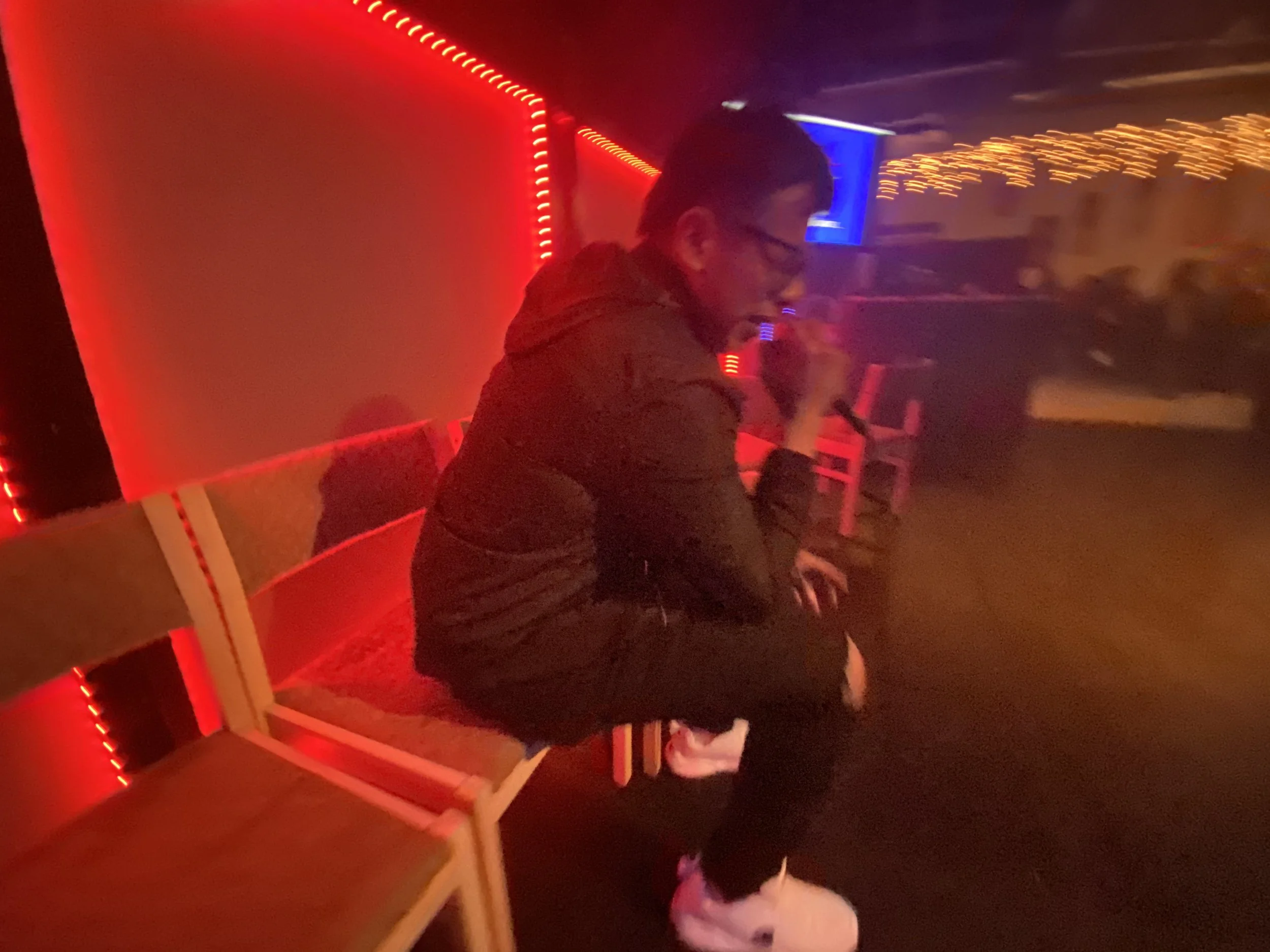 A man singing into a microphone in a dimly lit room with red lighting, sitting on a wooden bench, wearing glasses, a black jacket, and white shoes.