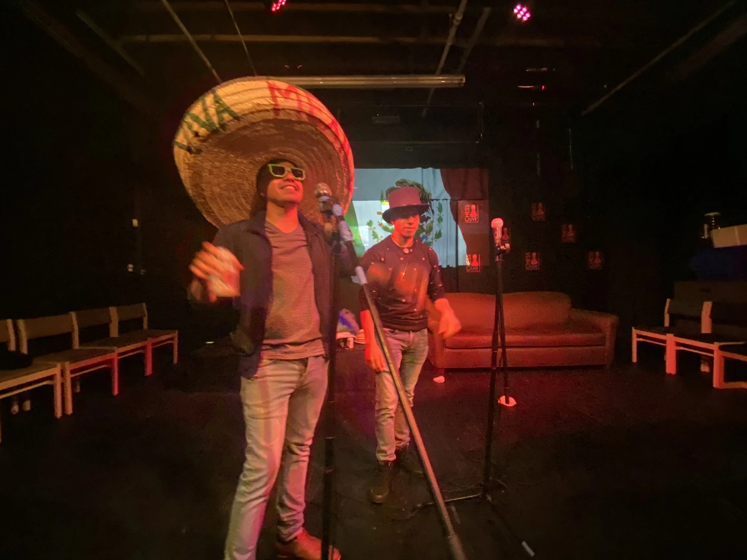 Two performers standing on a stage with microphones, wearing large sombreros, one with sunglasses, in a dimly-lit venue with chairs around and a background screen showing a colorful image.