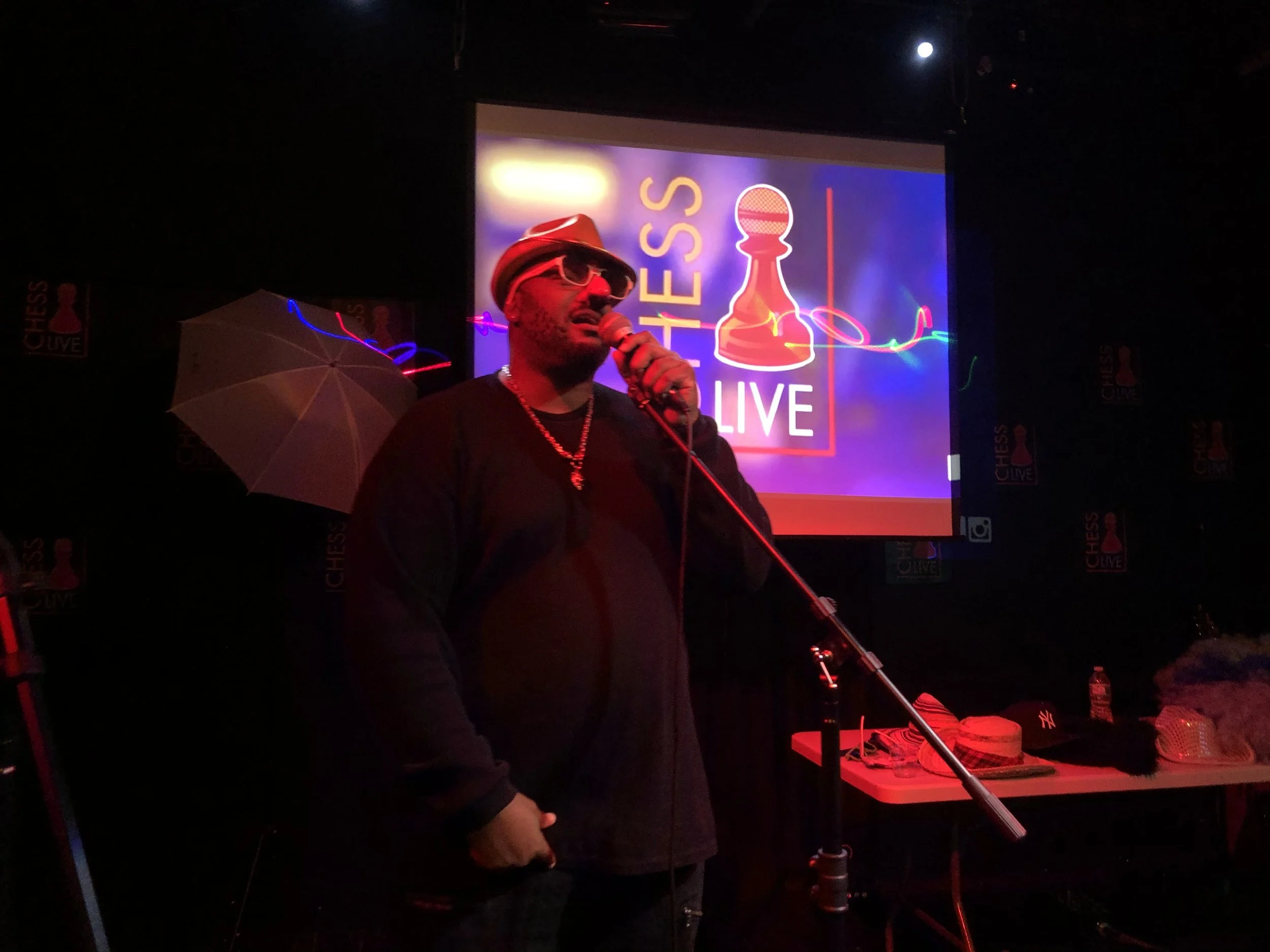 Man wearing white hat, sunglasses, black long-sleeve shirt, and gold chain, holding a microphone on stage with a screen behind him displaying the logo for Chess Live.