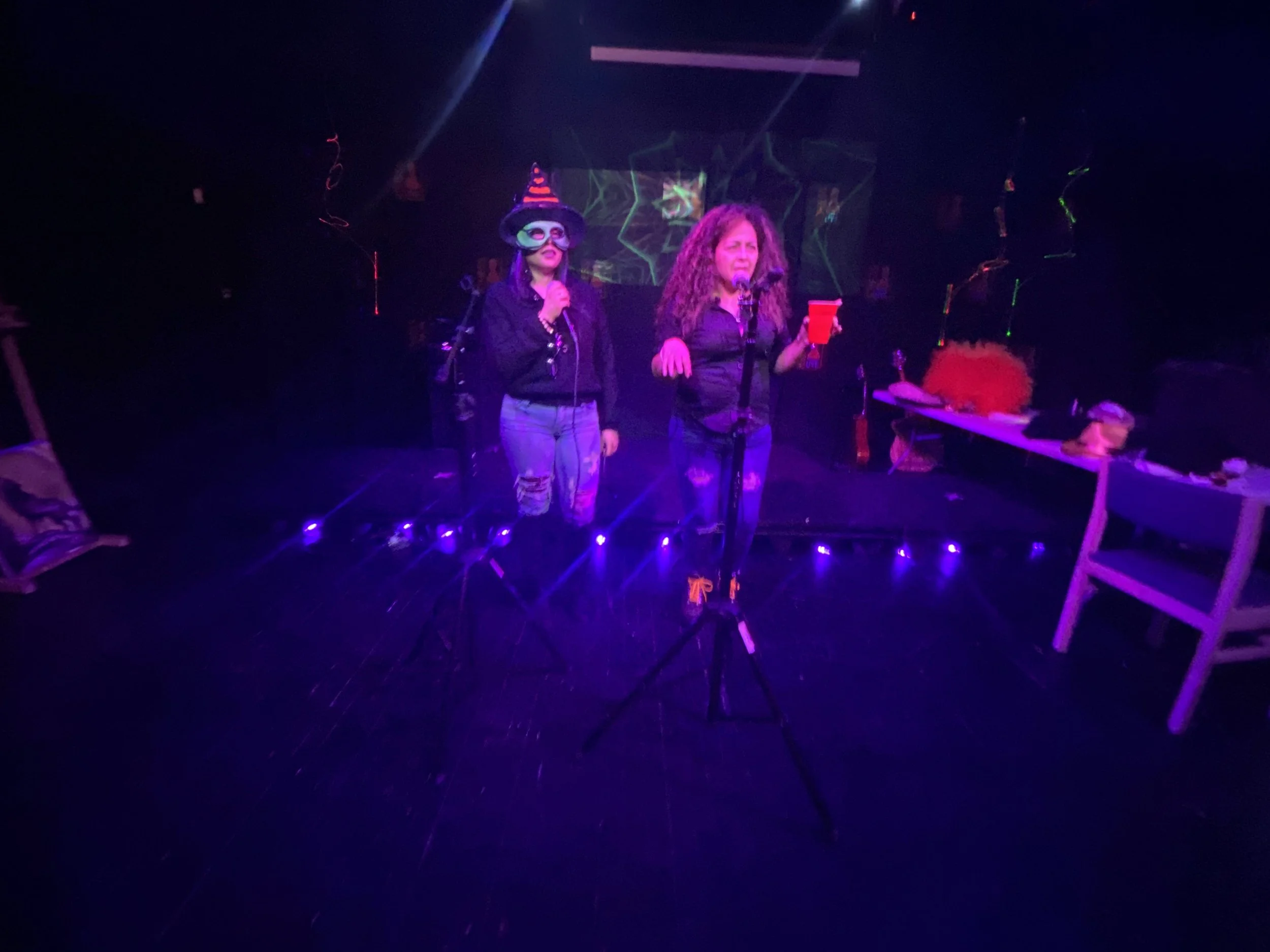 Two women stand on a dark stage, one with curly hair holding a phone and the other wearing a witch hat and mask, in a costume party setting with purple and green lighting.