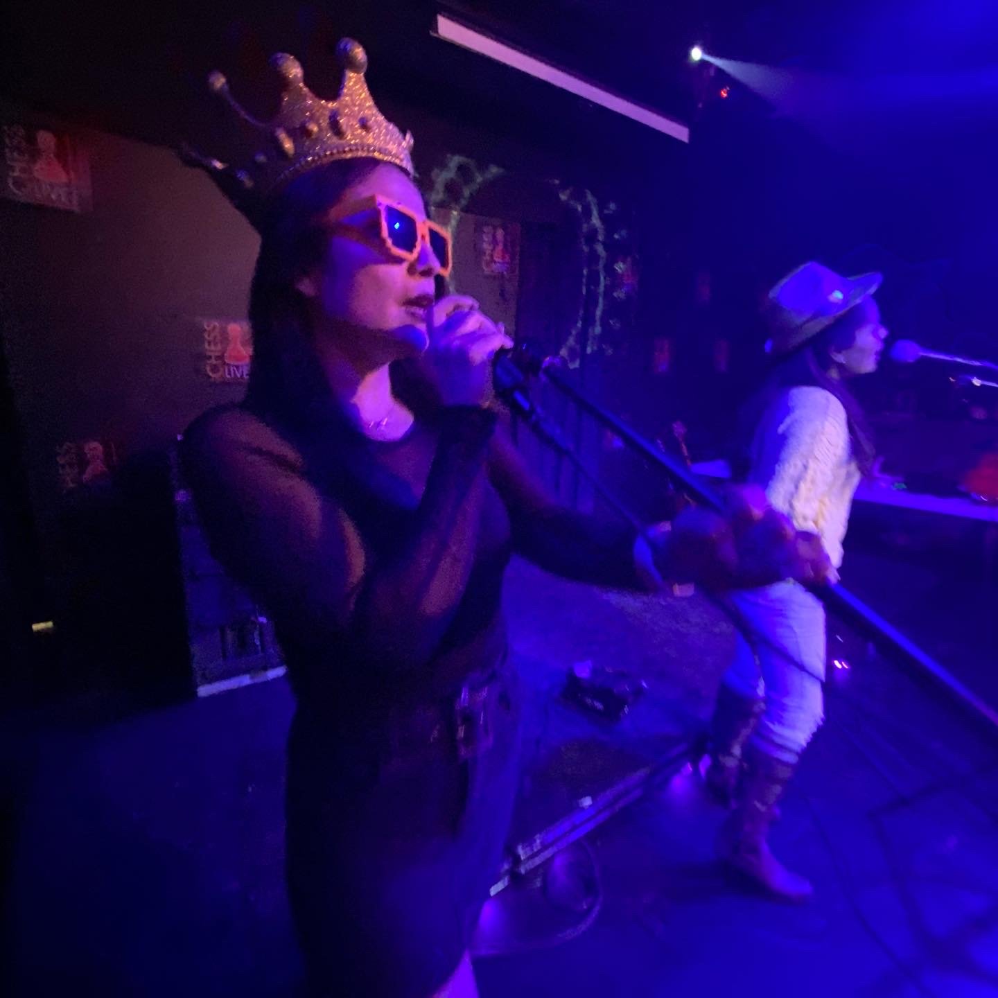 Two women performing on stage with microphones, styled in colorful accessories, under purple and blue stage lighting.