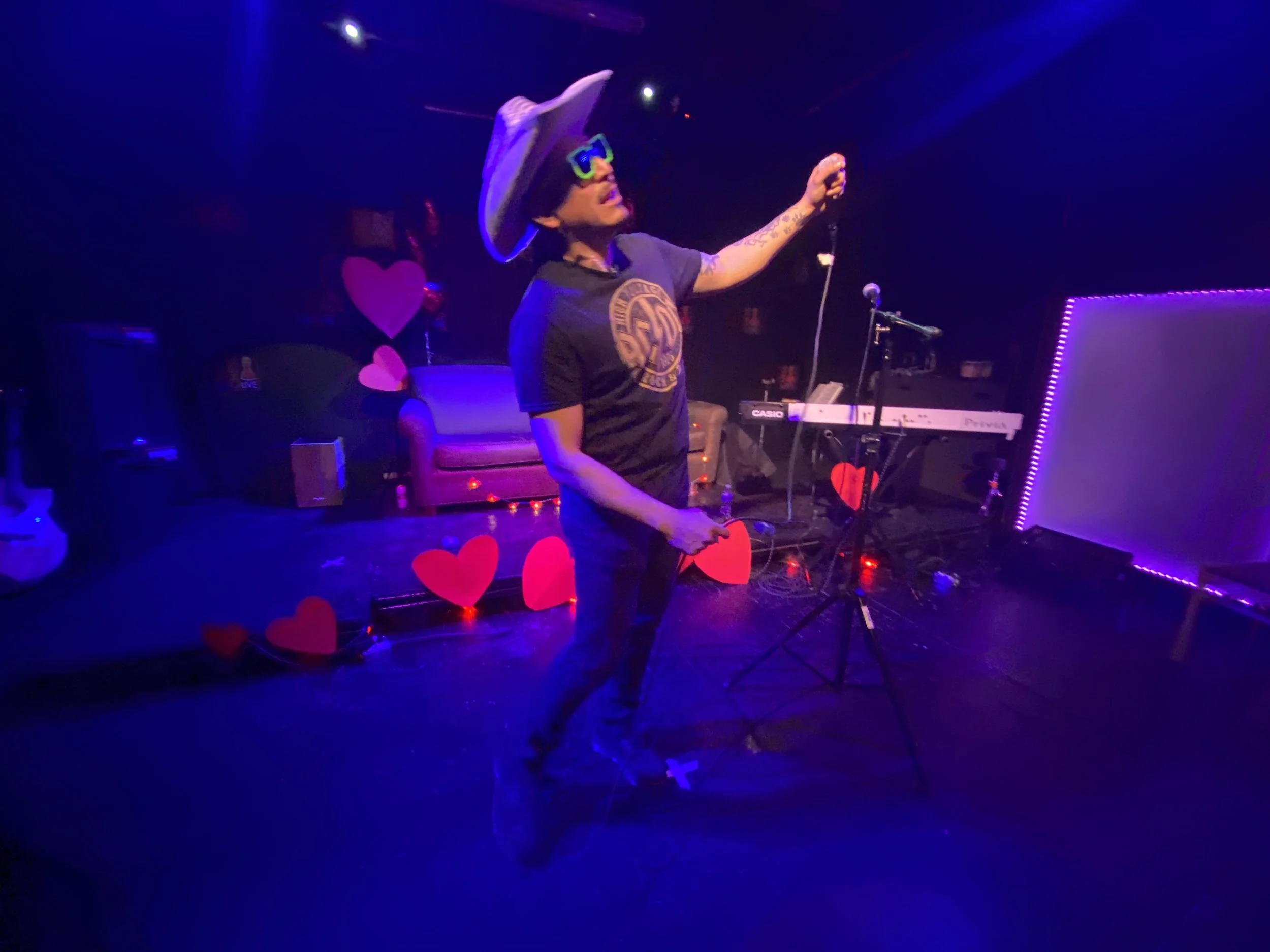 A performer on stage holding a microphone, wearing a large hat, green glasses, and a T-shirt with a circular design. The stage is decorated with red heart shapes, and there is electronic equipment including a keyboard and microphone stand visible.