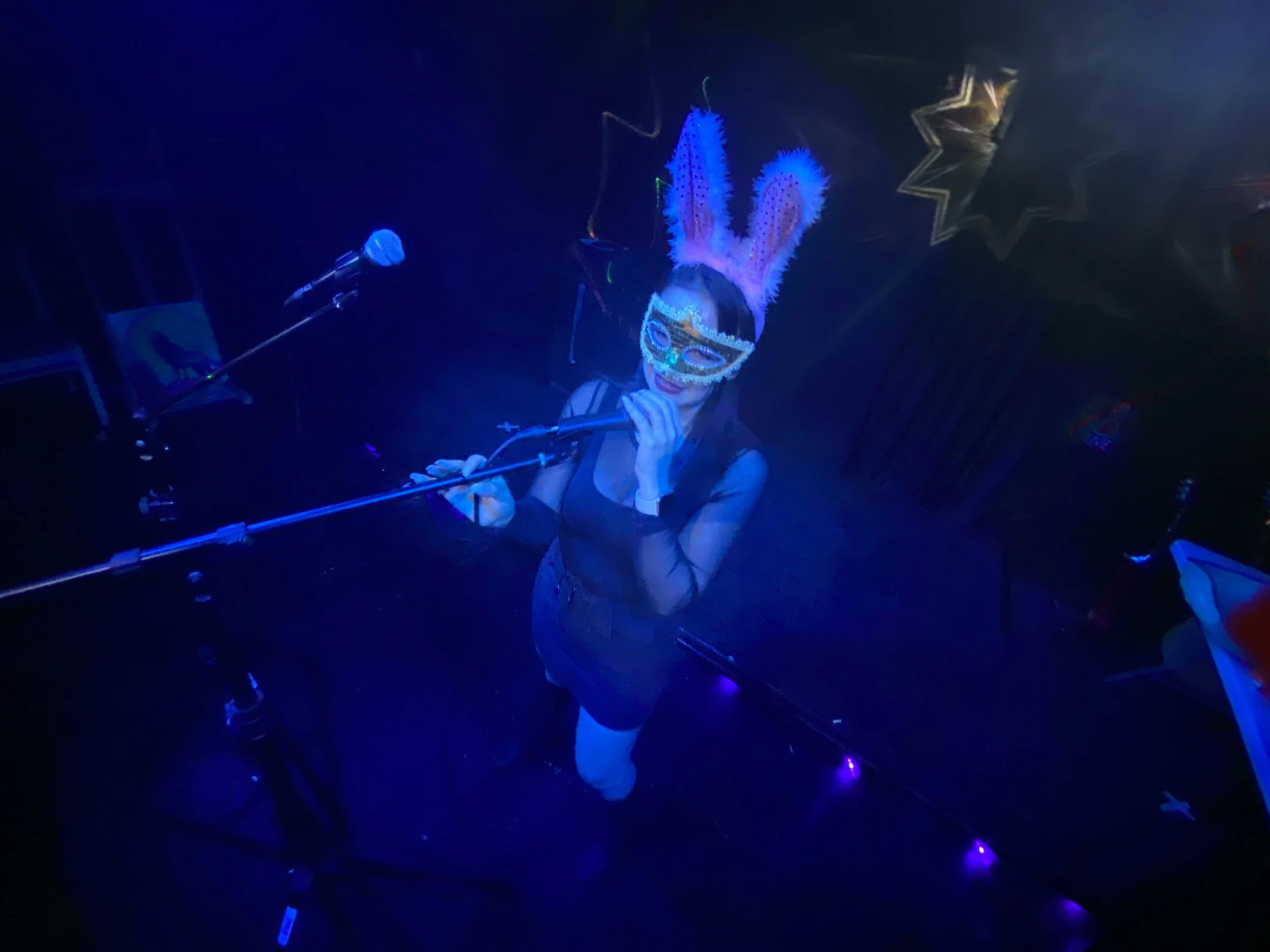 Woman wearing bunny ears and a decorative mask singing into a microphone on stage with blue lighting.
