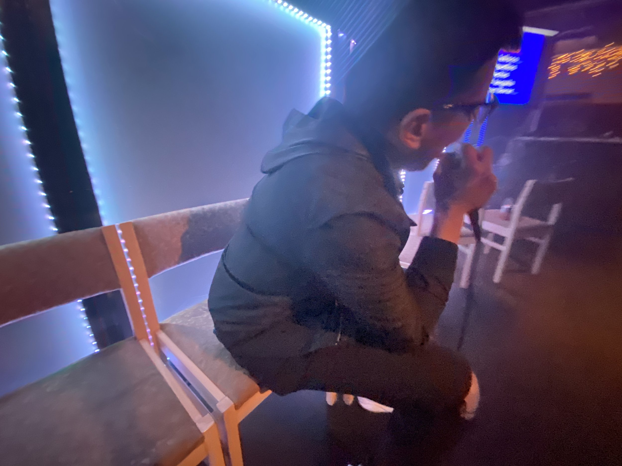 A man sitting on a bench in a dimly lit room, holding a microphone near his mouth, with neon blue and orange lights in the background.