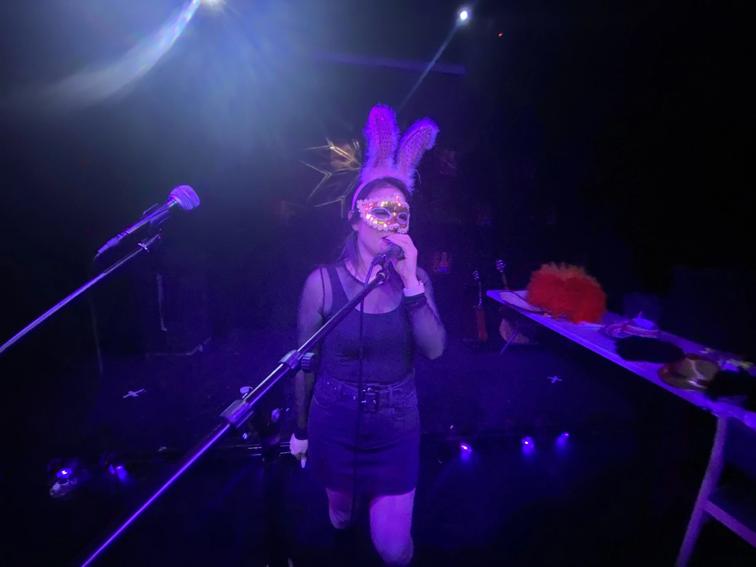 Person singing on stage with a bunny ears headband, wearing a colorful mask, and holding a microphone, illuminated by purple stage lights.