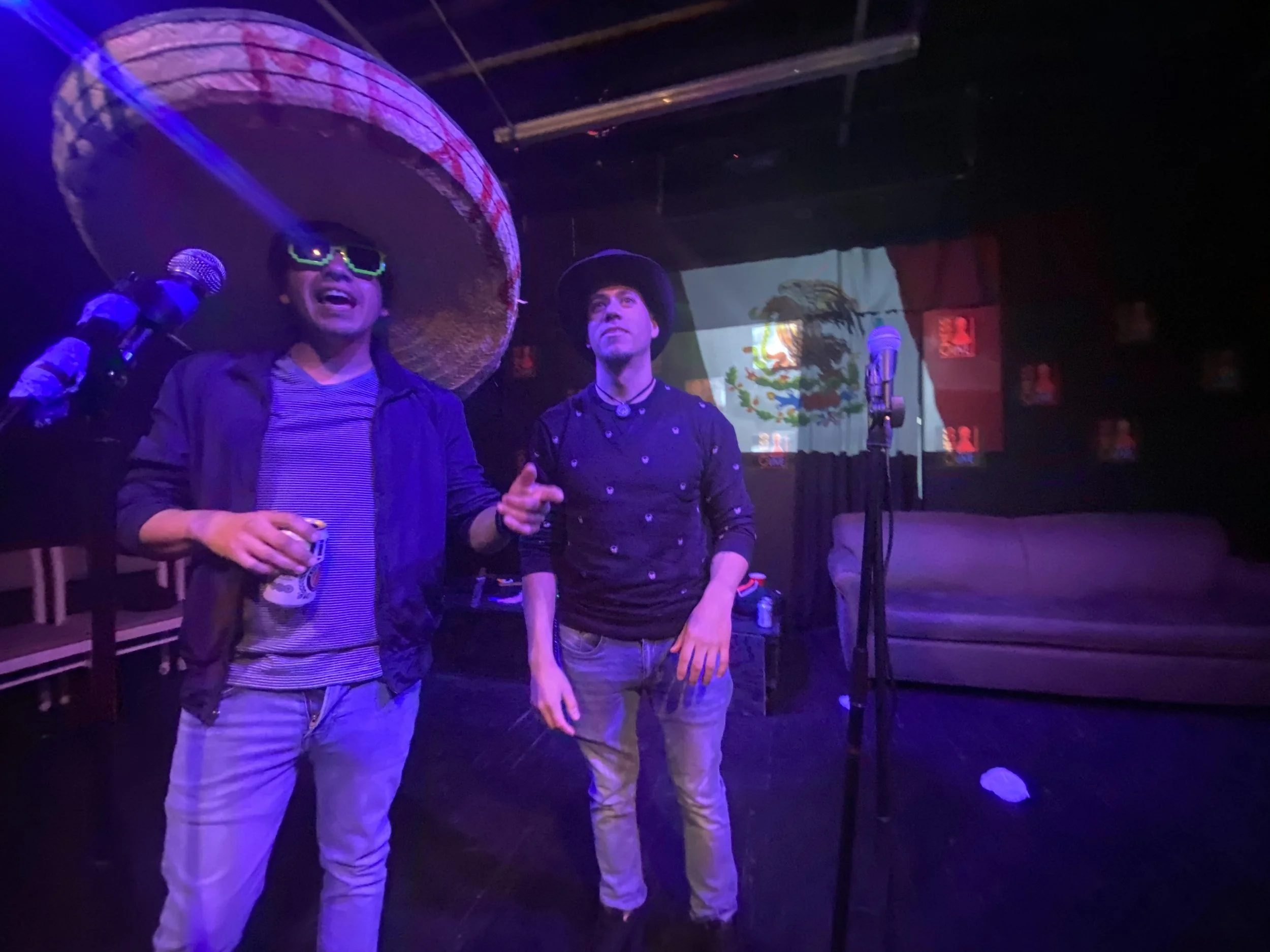 Two men with Mexican-themed costumes and accessories stand on a stage with a Mexican flag projection in the background. One wears a large sombrero and sunglasses, holding a drink, while the other wears a black hat, with microphones nearby, under purp