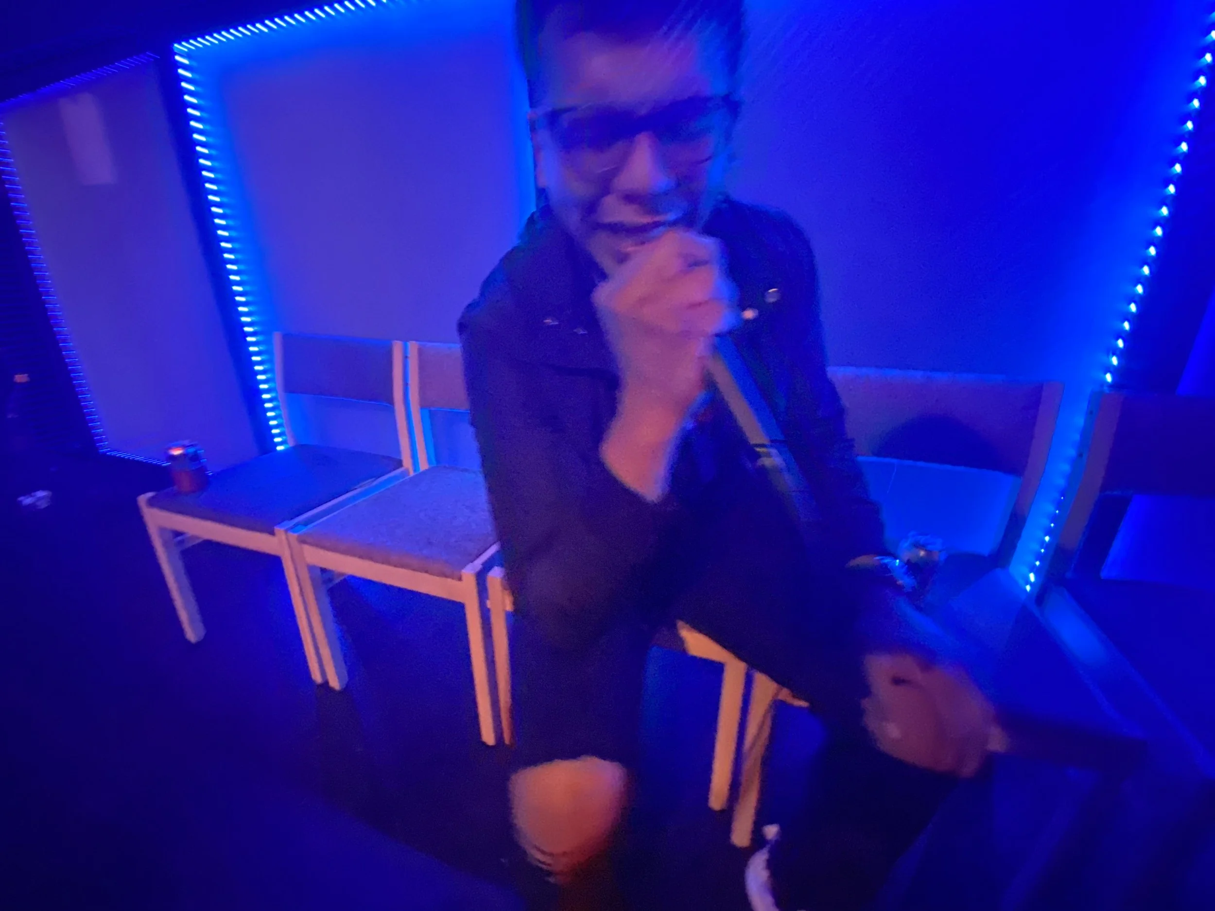 Person singing karaoke in a dimly lit room with blue LED lighting, holding a microphone and sitting on a chair.