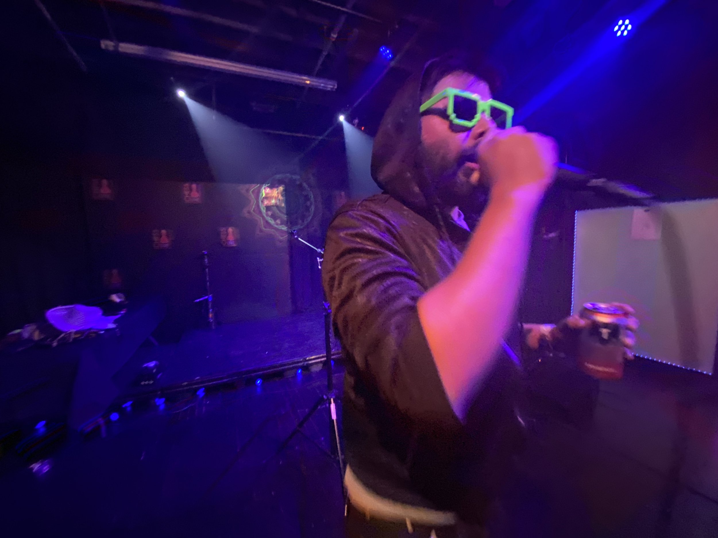 A man wearing green, pixelated sunglasses and a hoodie is singing or speaking into a microphone on stage at a nightclub or concert venue, with colorful lights and a dark background.