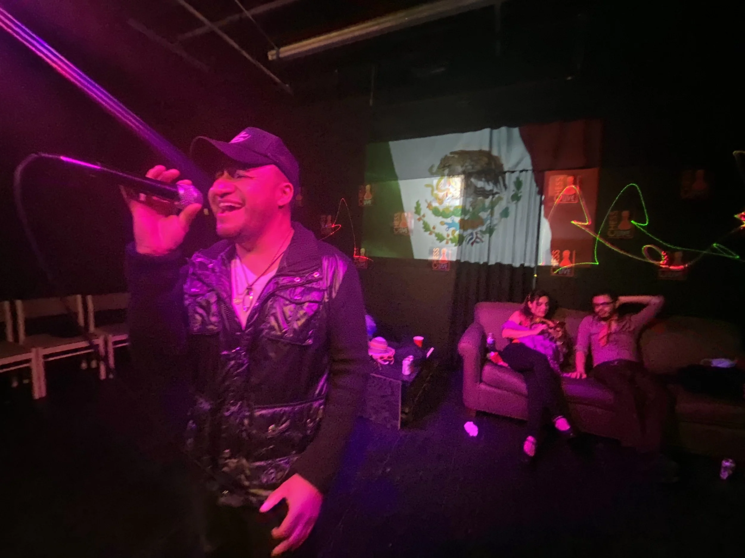 A man singing with a microphone in a nightclub with a Mexican flag projected in the background, and two people sitting on a sofa.