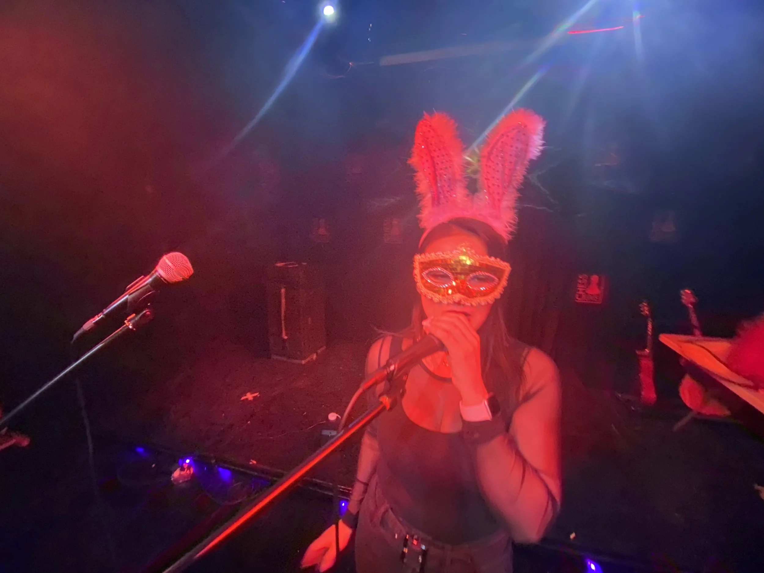 A woman wearing pink bunny ears and decorative glasses singing into a microphone in a dimly lit venue with colorful stage lights.
