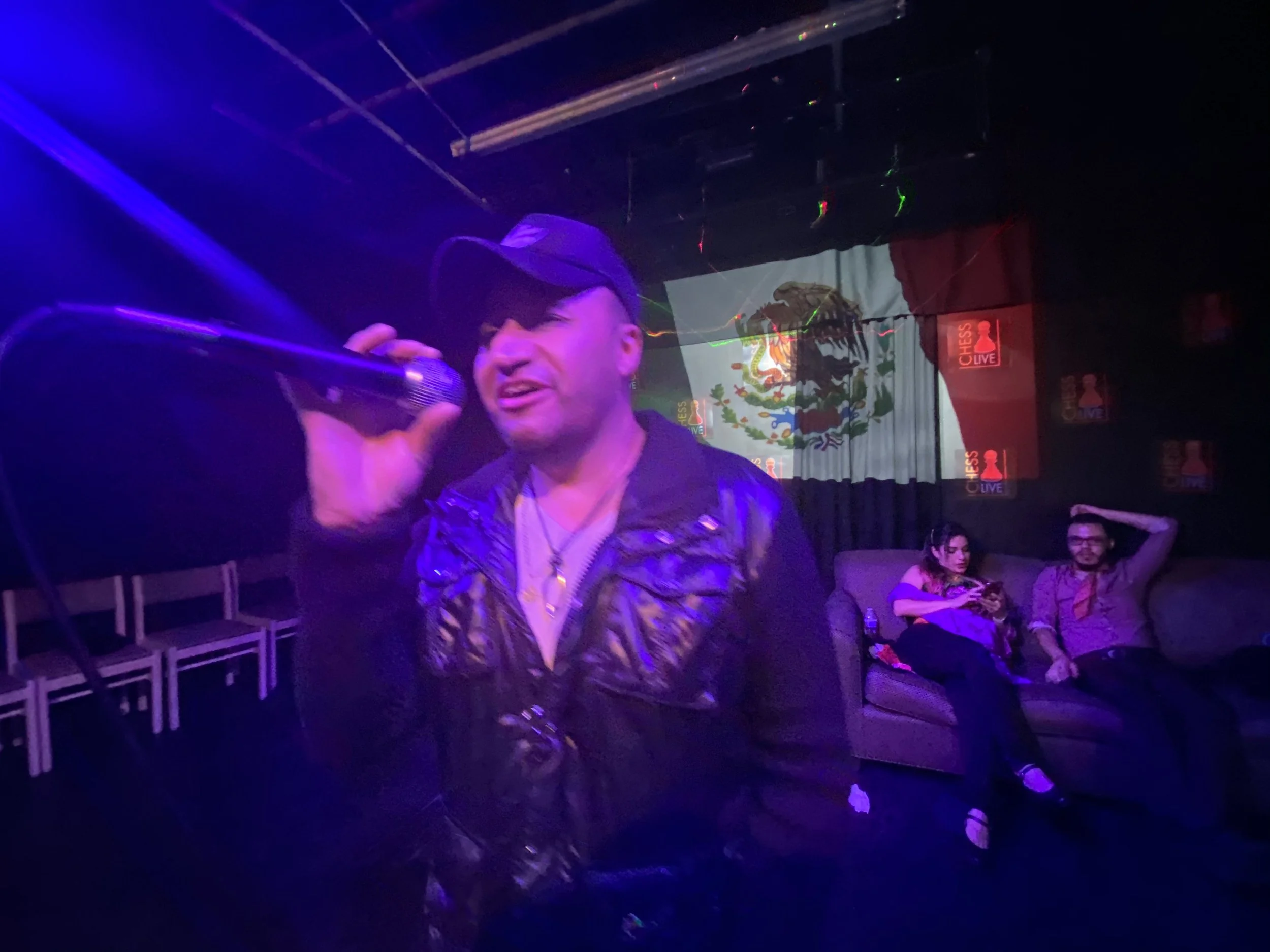 A man singing into a microphone at a karaoke bar with Mexican flags and Chess Live banners on the wall behind him. Two people sitting on a gray couch in the background, one looking at a phone and the other resting with a hand on his head.