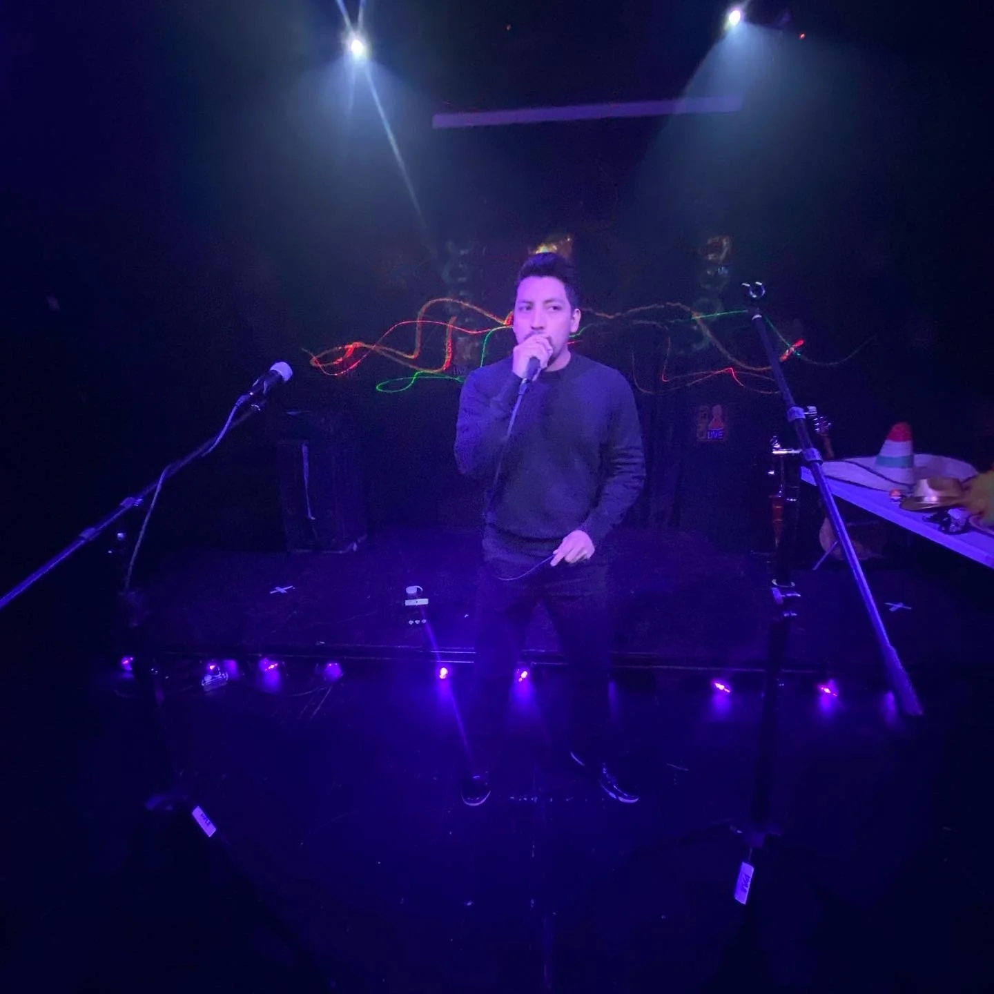 A man standing on a stage with purple lighting, holding a microphone, with two microphones on stands in front of him. There are colorful light streaks behind him, and some equipment and items on the right side of the stage, including a traffic cone a