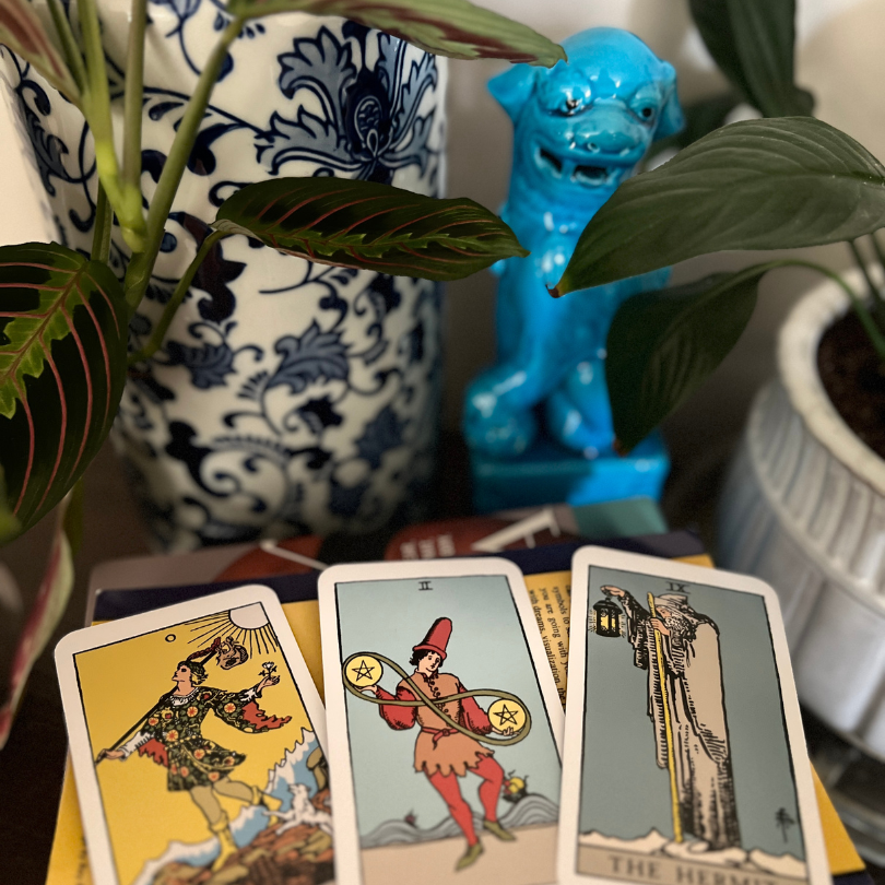 Three Rider-Waite tarot cards (The Fool, Two of Pentacles, and The Hermit) arranged on books beside houseplants, illustrating the beginning of a personal tarot practice.