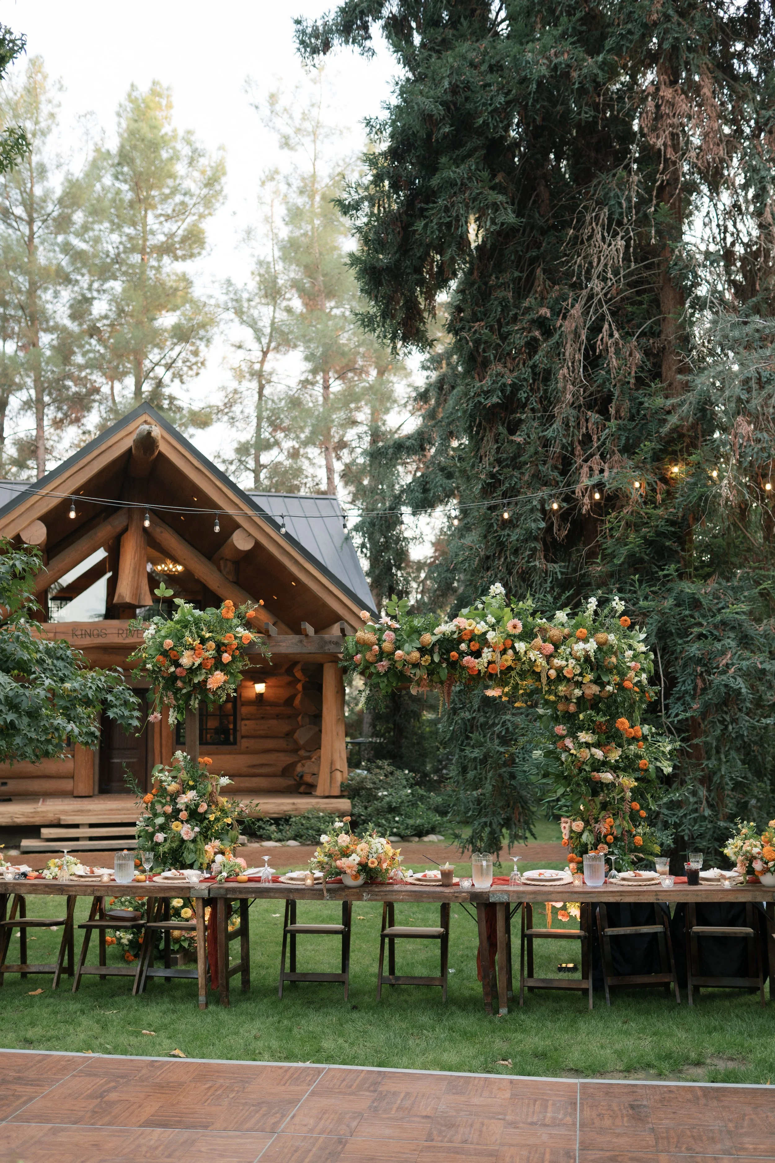 Outdoor event setup with a long wooden table decorated with flower arrangements, plates, and glasses, set in front of a log cabin with string lights and surrounded by tall trees.