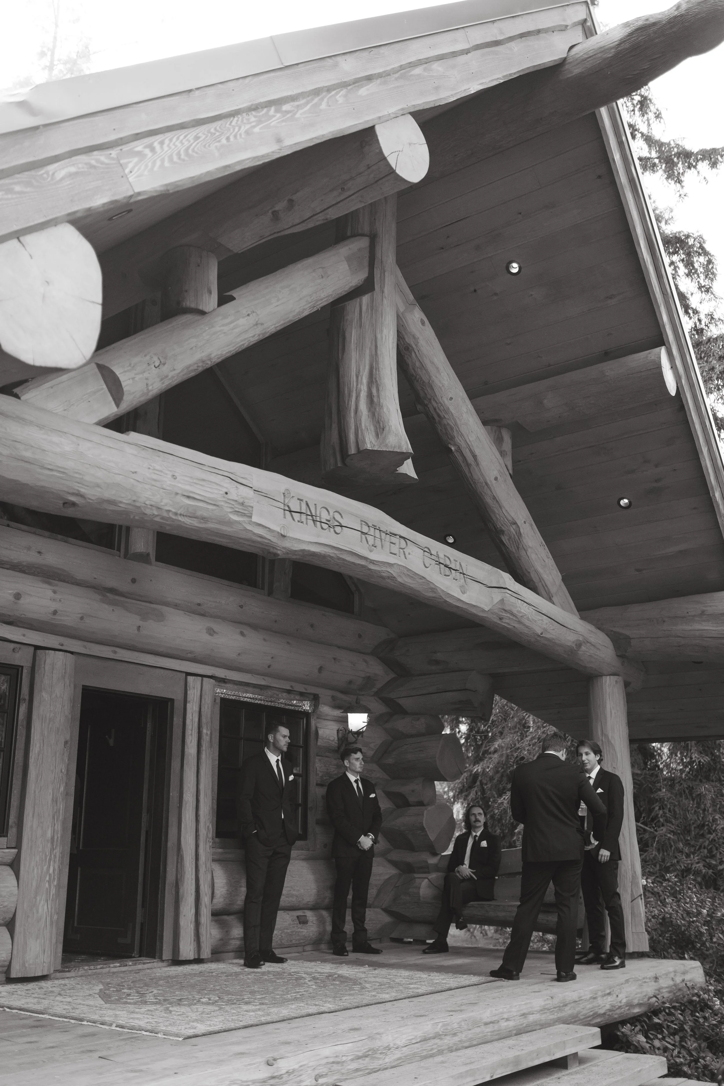 A group of five men dressed in suits standing and sitting on a wooden porch of a log cabin-style building with a sign that reads "KINGS RIVER CABIN". The porch has a rustic design with logs and beams.