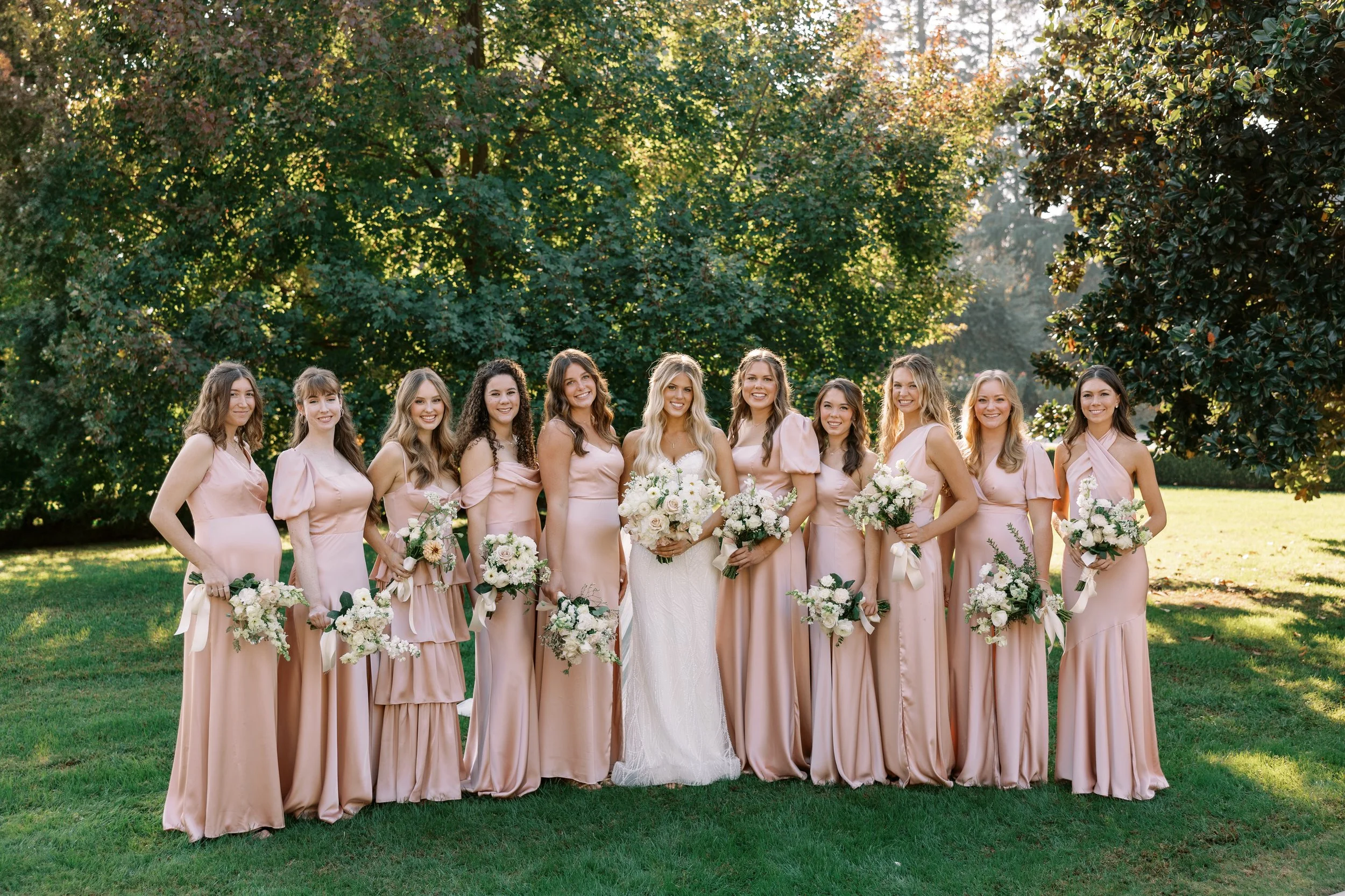 A bride in a white wedding dress with ten bridesmaids in pale pink dresses holding bouquets, standing on a grassy field with green trees in the background.