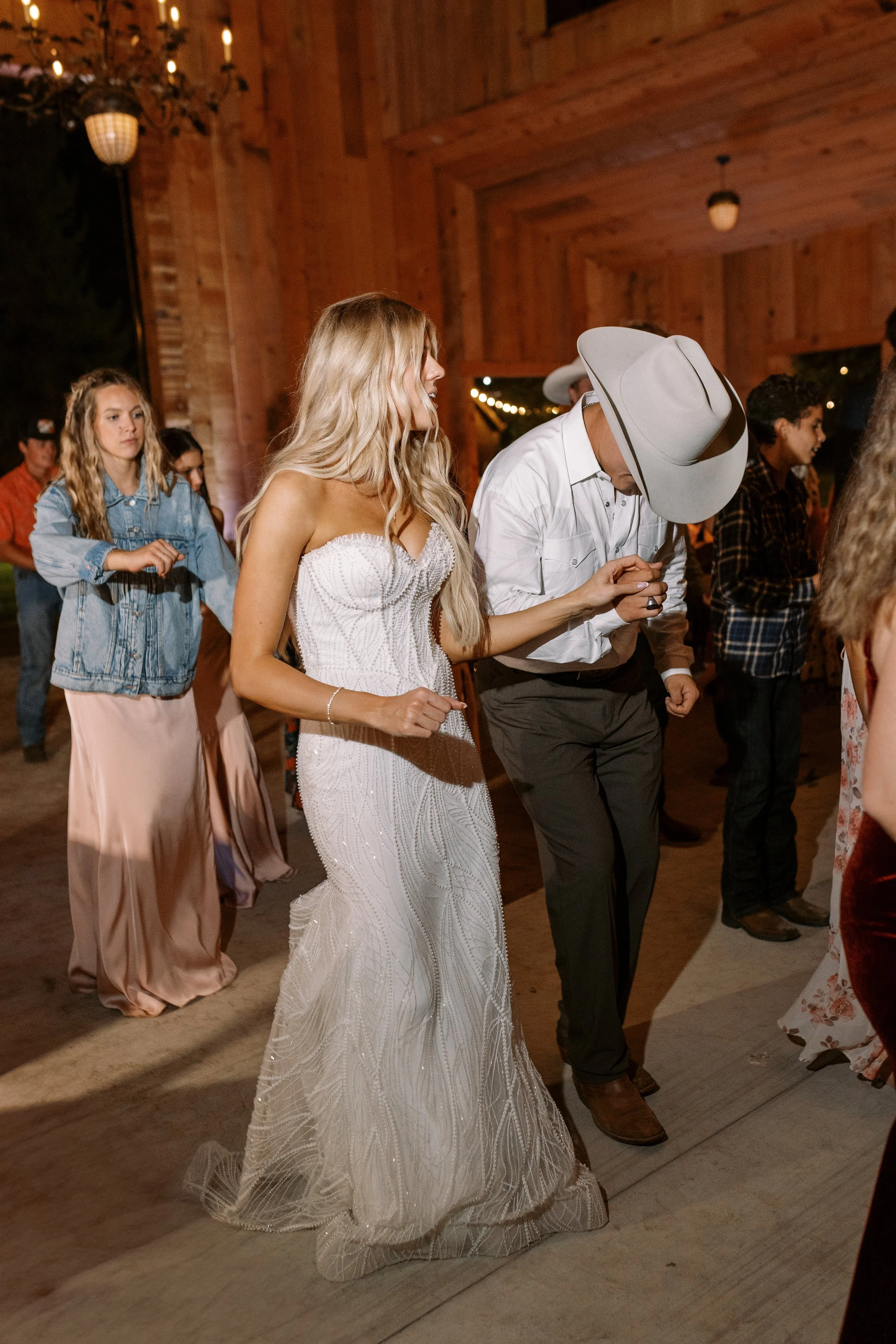 A bride in a white wedding dress and a groom in a white shirt with a large gray cowboy hat dancing at a rustic wooden wedding reception.