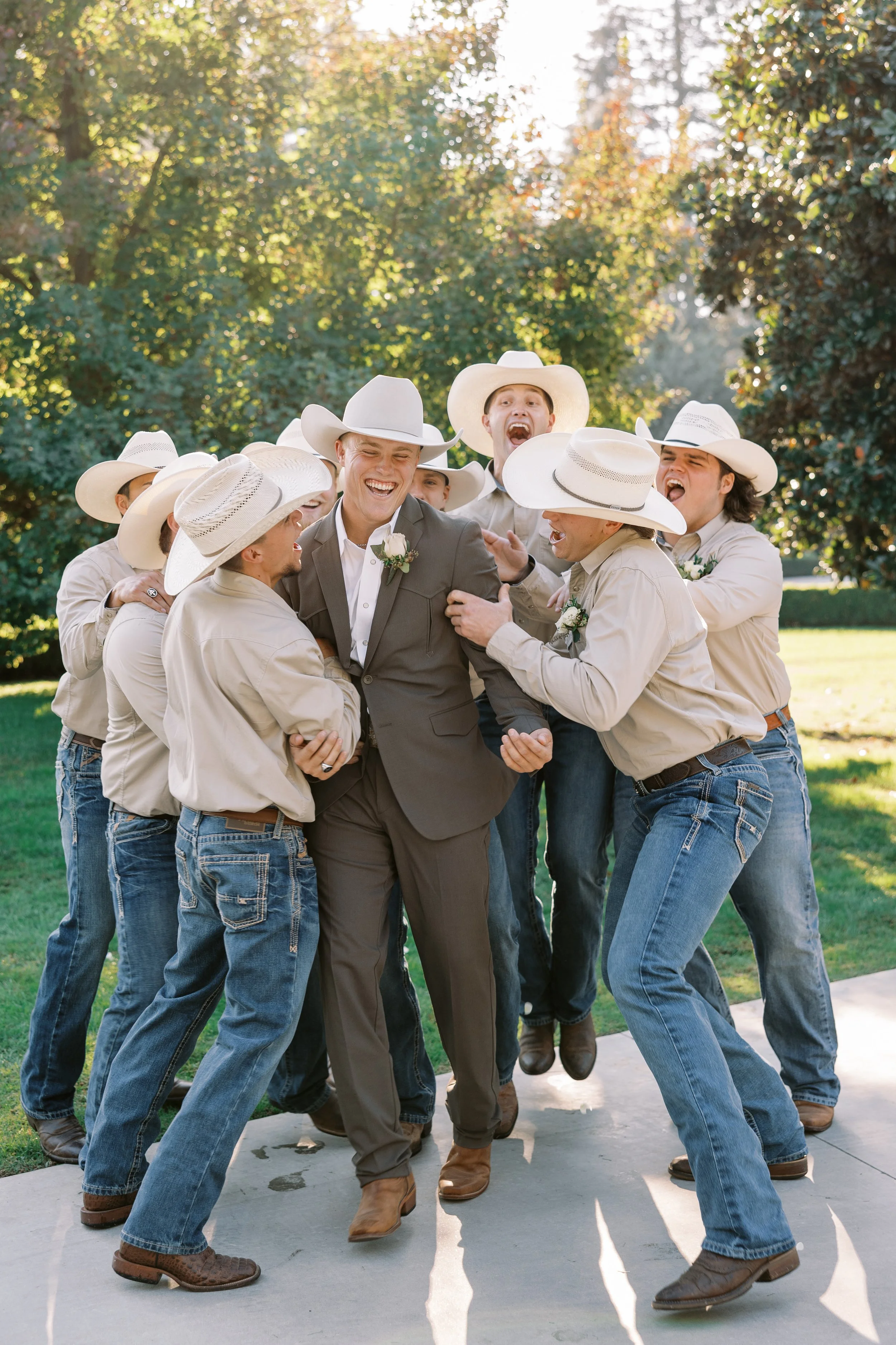 Group of men, some wearing cowboy hats, laughing and playfully grabbing a man in a suit outdoors on a sunny day.