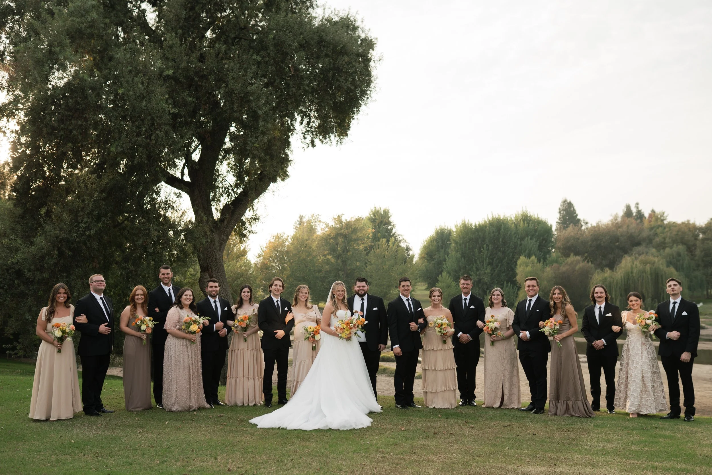 Group of wedding party members, including bride in white gown, surrounded by bridesmaids and groomsmen, standing outdoors on grass with trees in the background