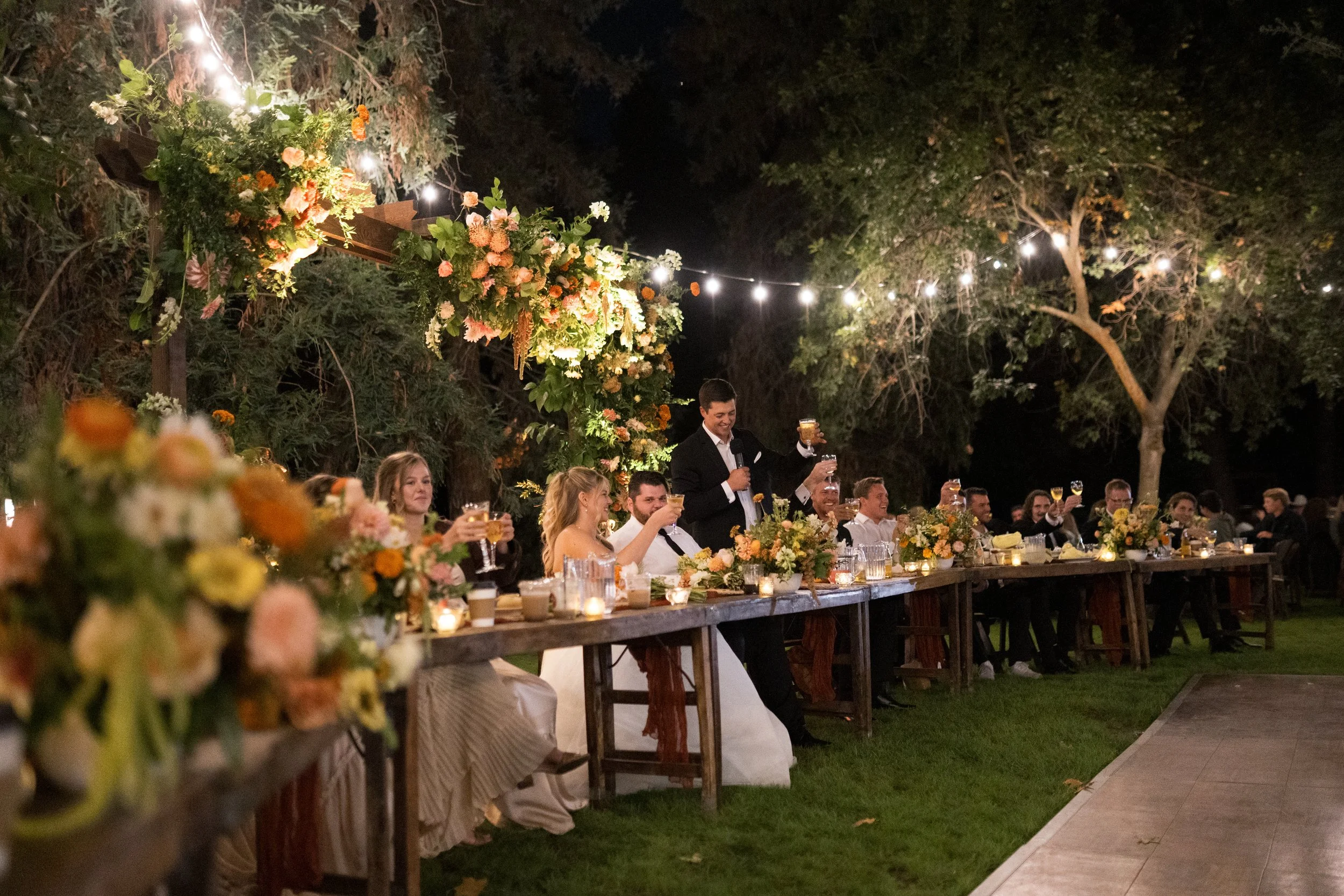 Outdoor wedding reception at night with long table decorated with flowers and candles, guests raising glasses, groom giving speech, string lights overhead, surrounded by trees.