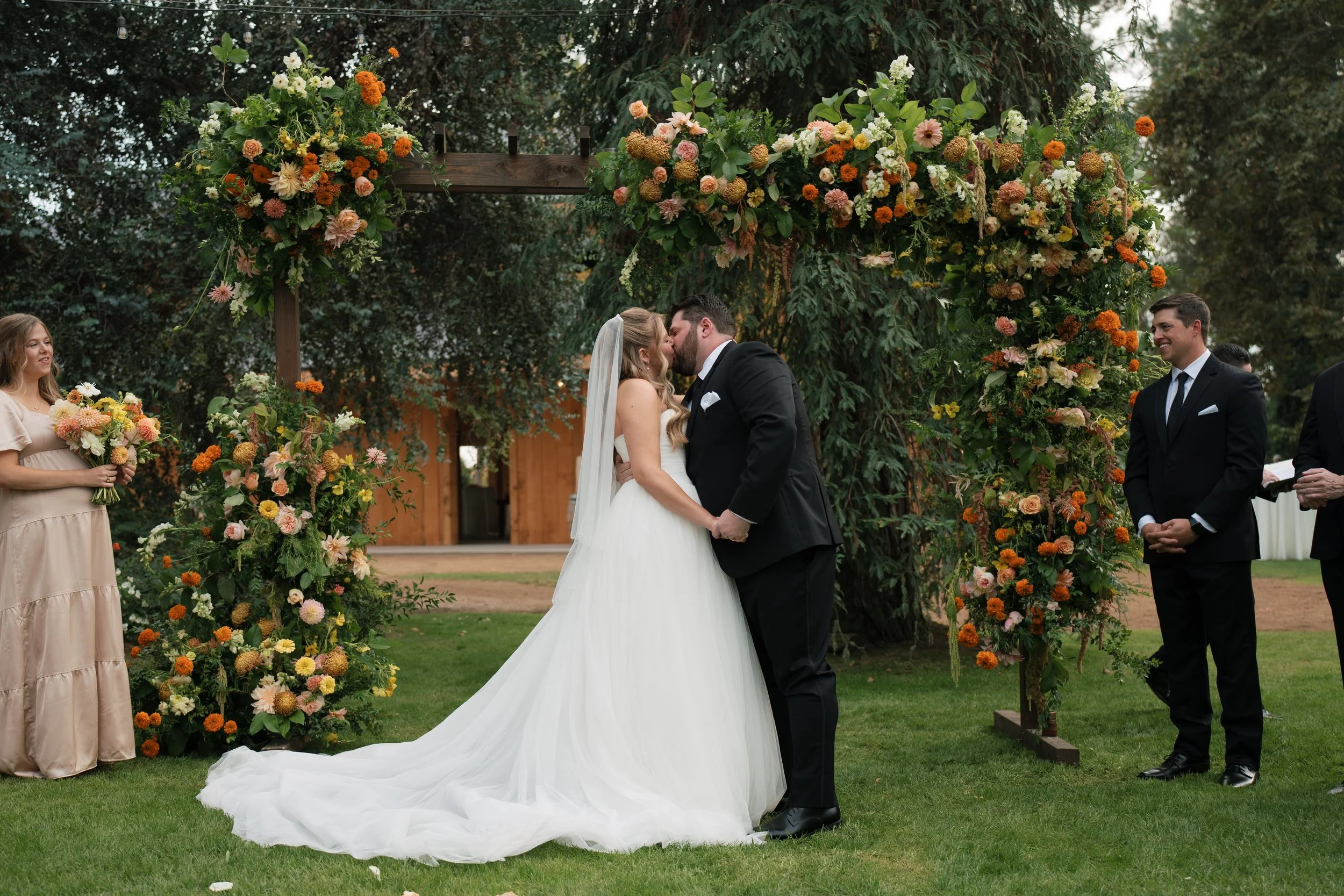 A bride and groom kiss during their outdoor wedding ceremony under a floral archway, with bridesmaid holding a bouquet on the left and groomsman watching on the right.