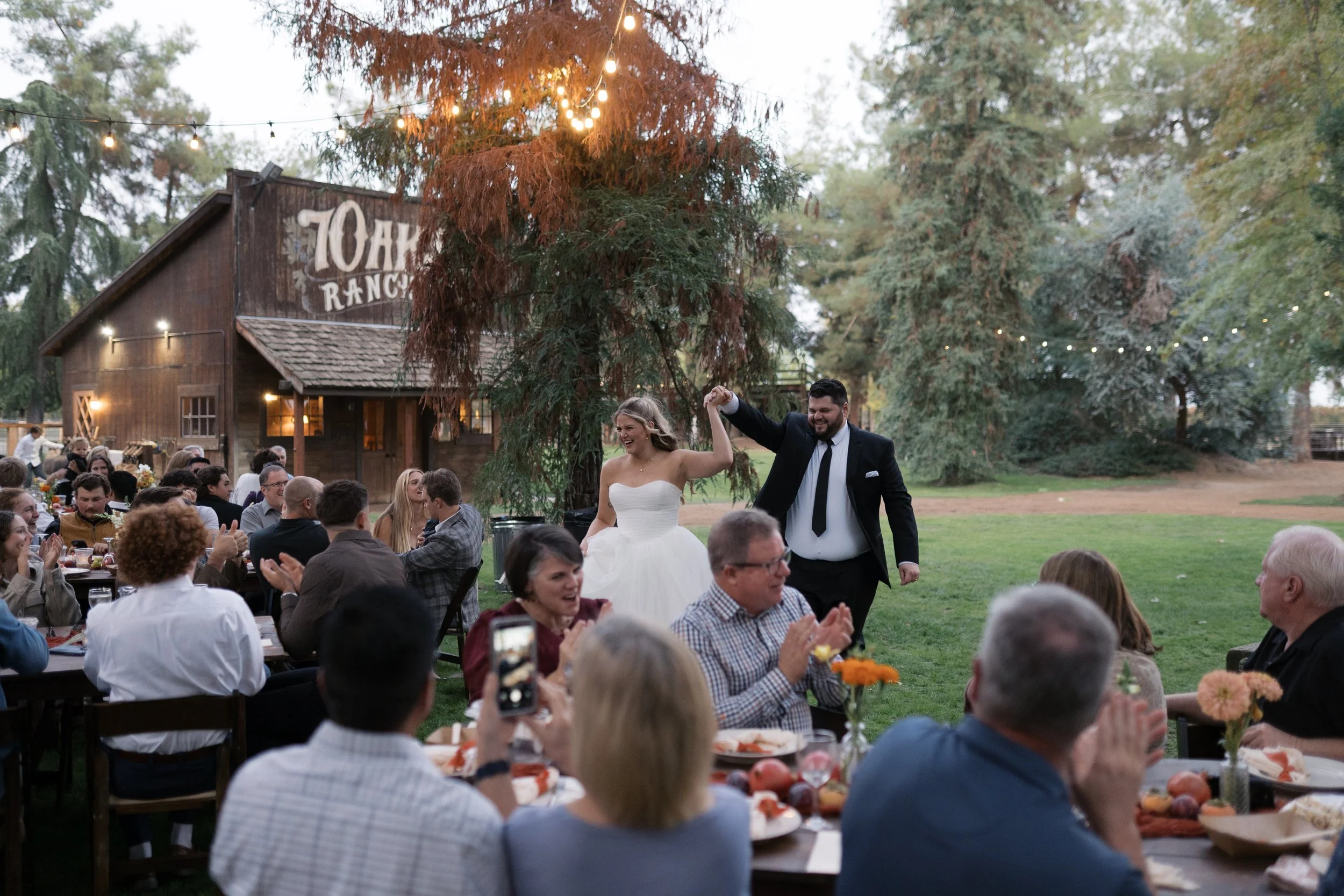 A bride and groom dancing and smiling at an outdoor wedding reception with guests seated at long tables, set against a rustic barn with string lights and large trees.