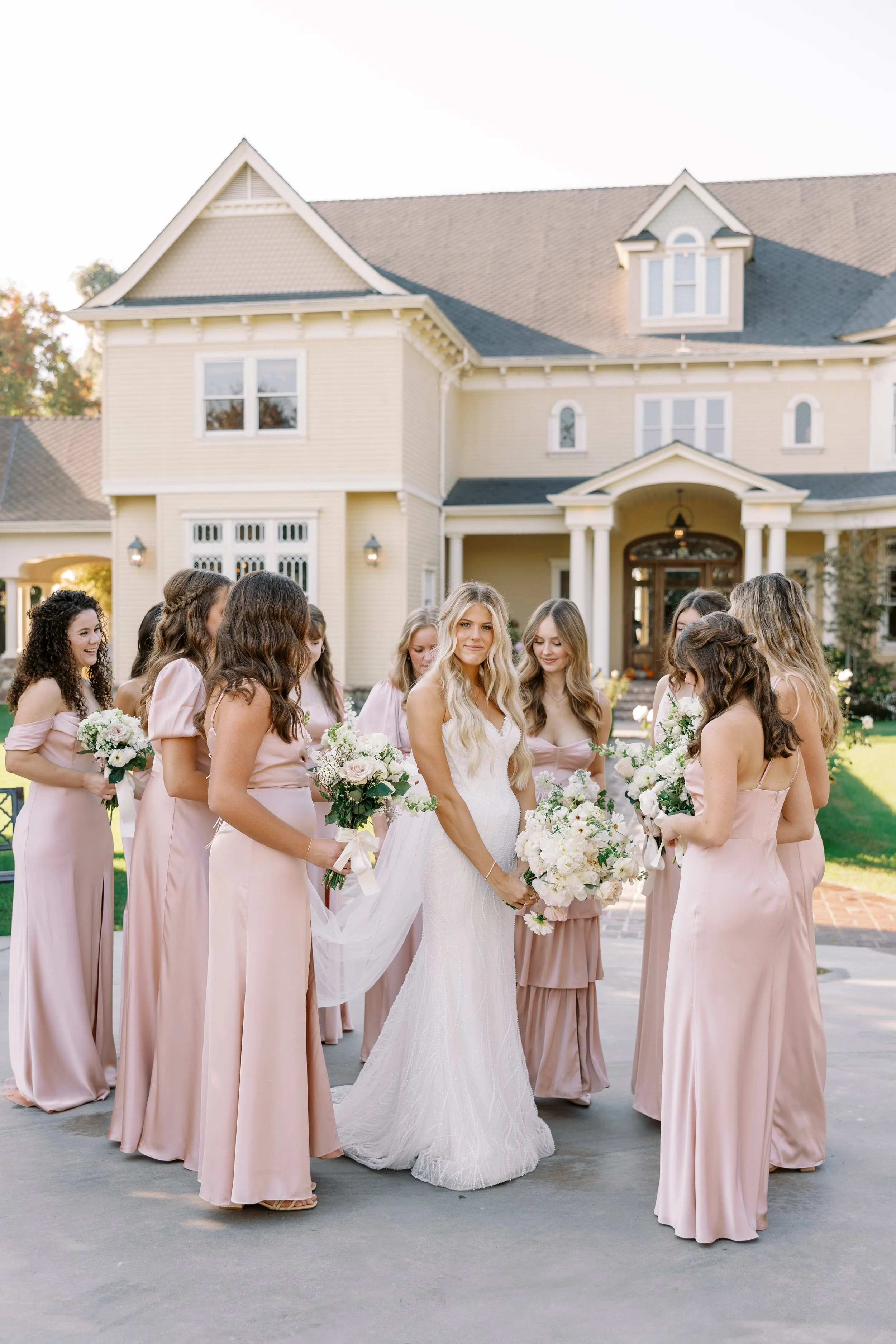 A bride and her bridesmaids standing outside in front of a large house, dressed in wedding attire, with flowers in hand.
