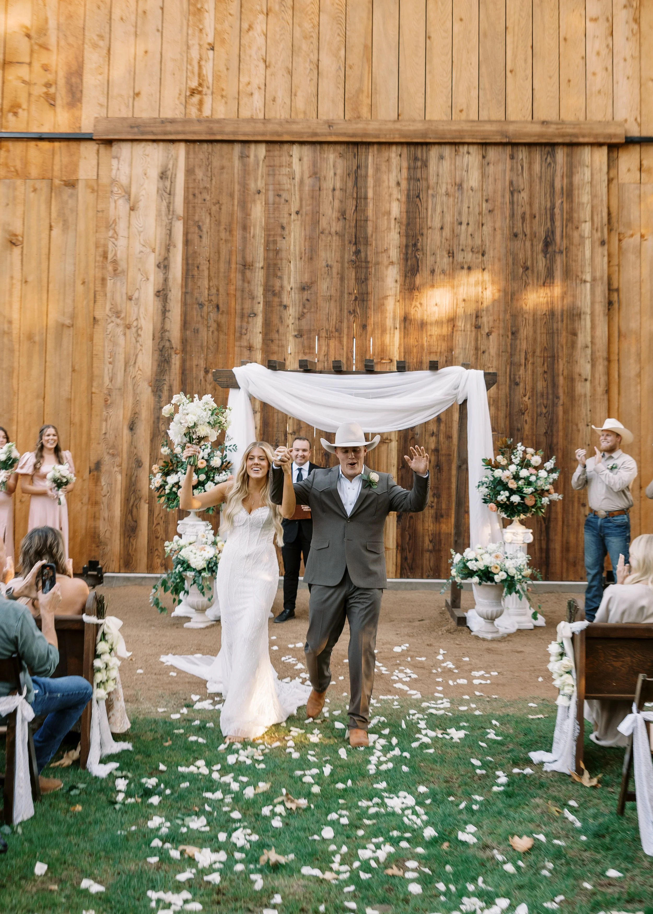 A bride and groom running down an aisle during a wedding ceremony in a rustic barn with wood walls, surrounded by flower arrangements and wedding guests.