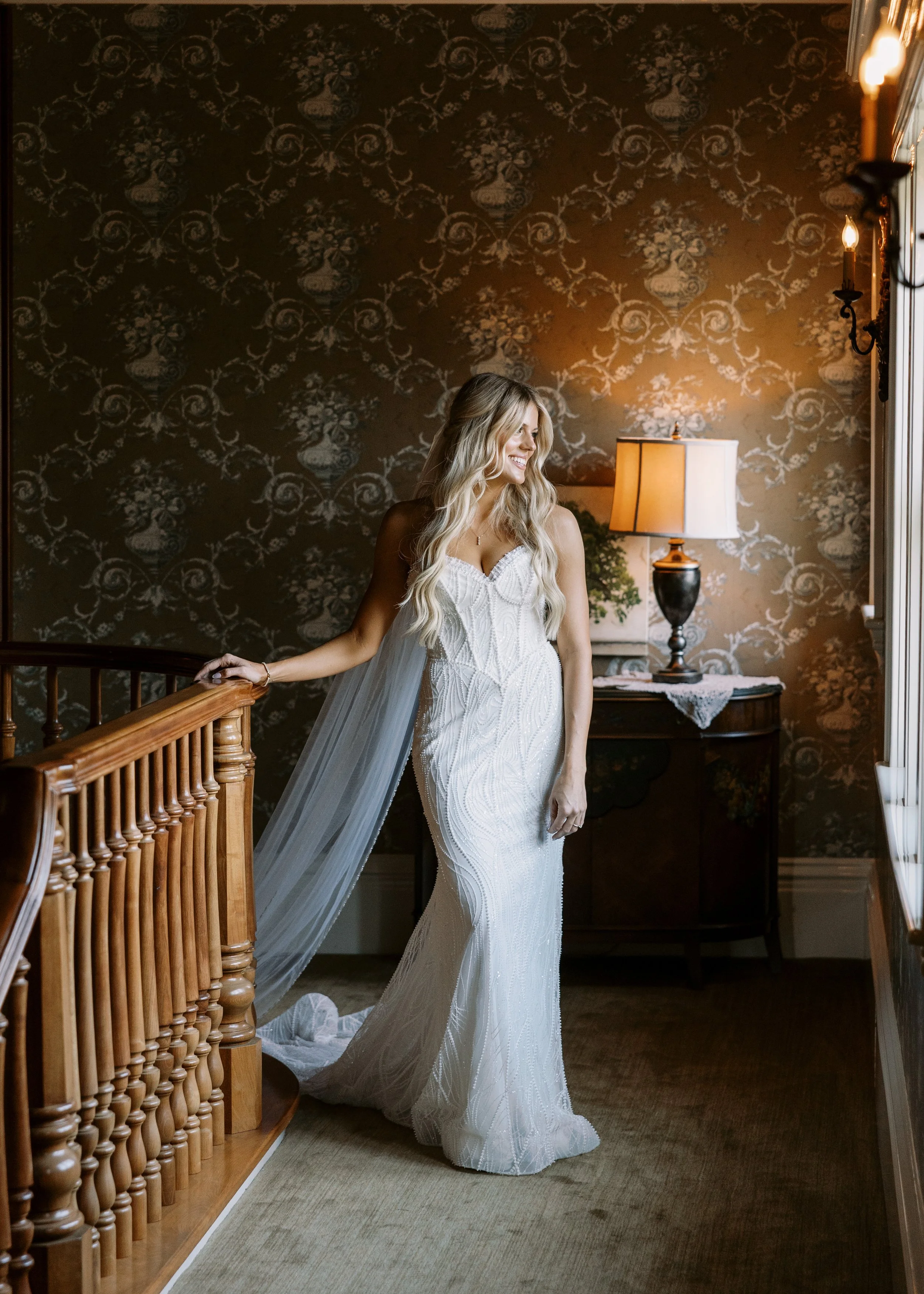 A bride in a white wedding gown standing on a staircase, smiling and looking away from the camera in a warmly lit room with floral wallpaper, a table with a lamp, and a window with natural light.