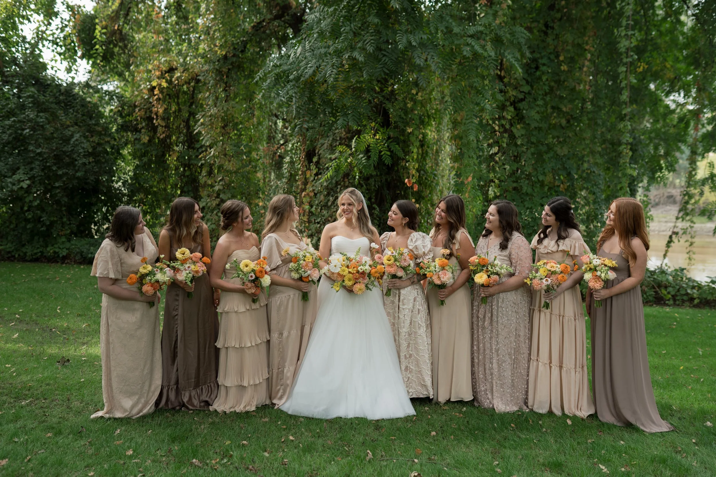 Group of women in beige dresses, standing on grass near trees, at a wedding, holding bouquets of orange, yellow, and white flowers, laughing and smiling.