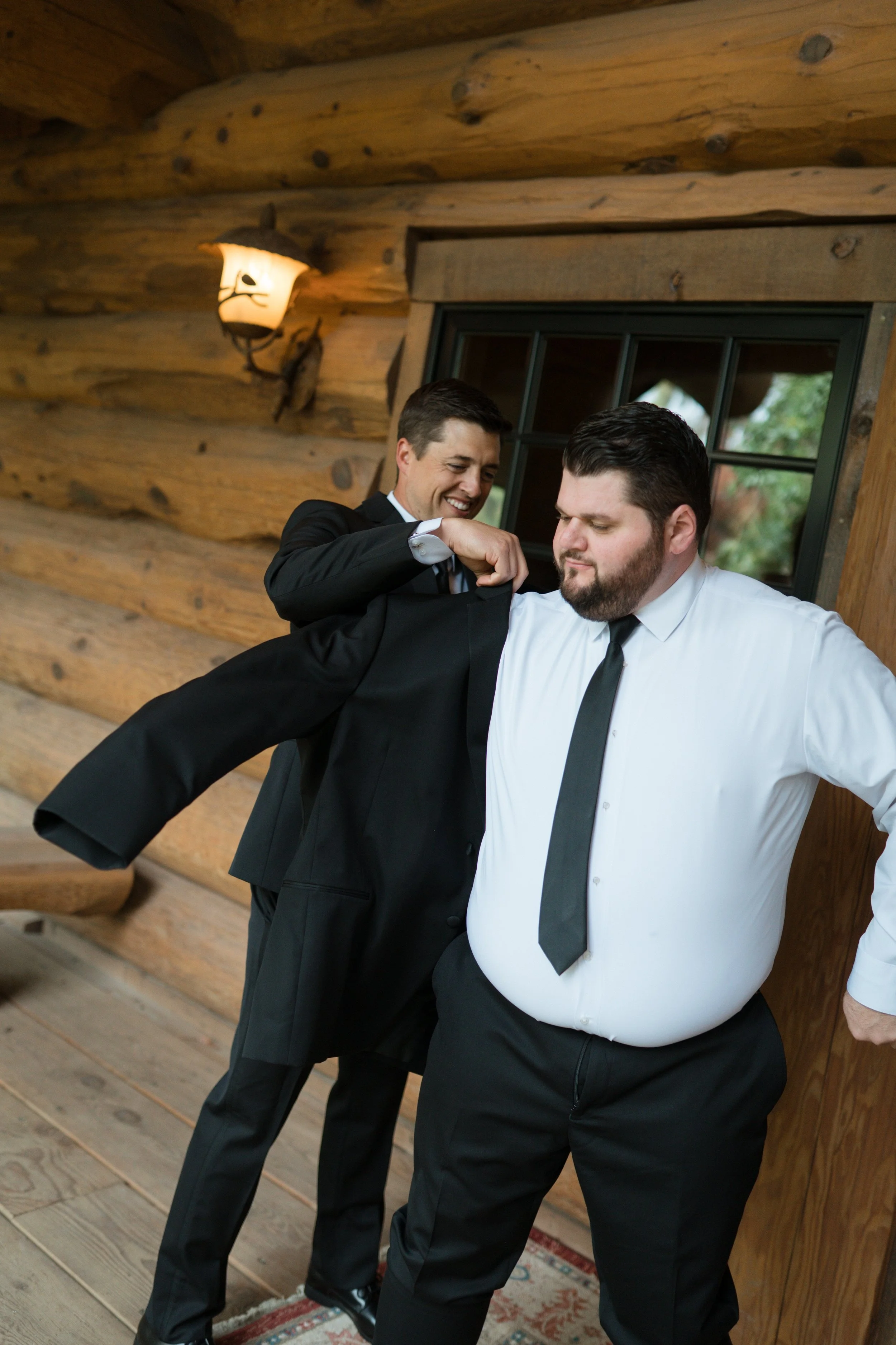 Two men in formal suits, one helping the other with his jacket inside a rustic wooden room.