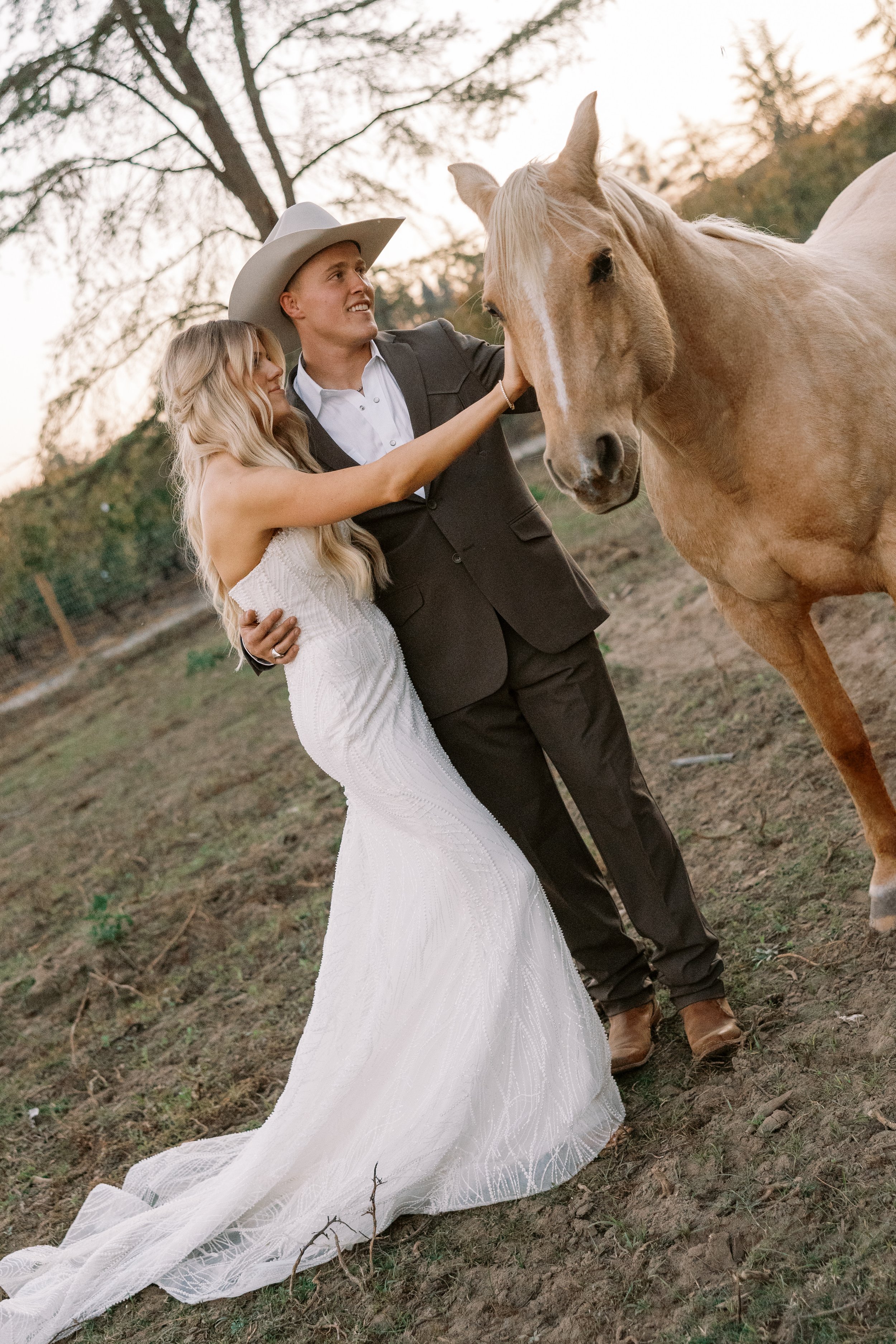 A bride and groom are outdoors, with the bride holding the groom as they pet a light tan horse, during sunset.