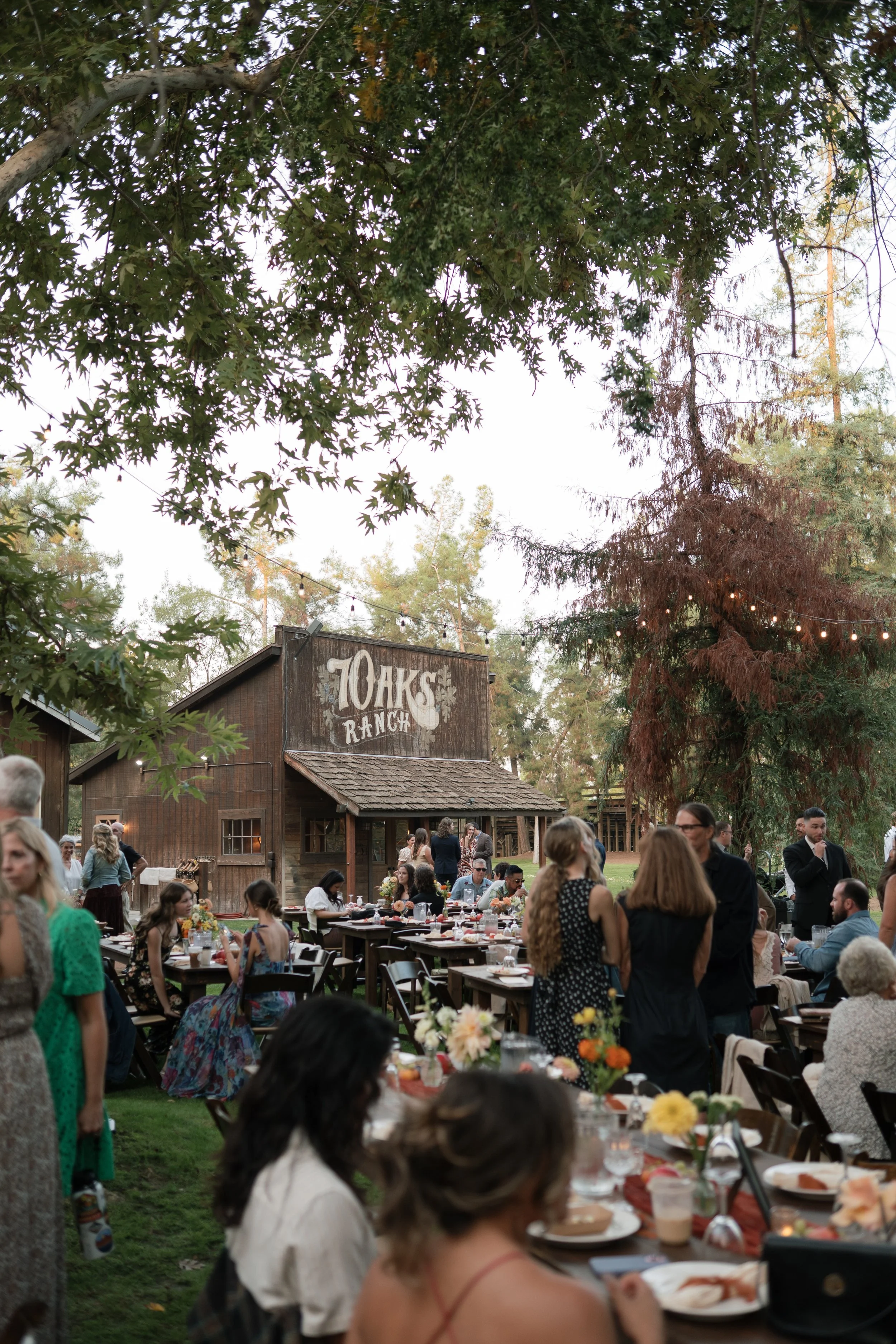 People gathered outdoors at a rustic wedding or event at Oaks Ranch, with tables decorated with flowers, in front of a wooden barn with a sign that reads 'Oaks Ranch'.