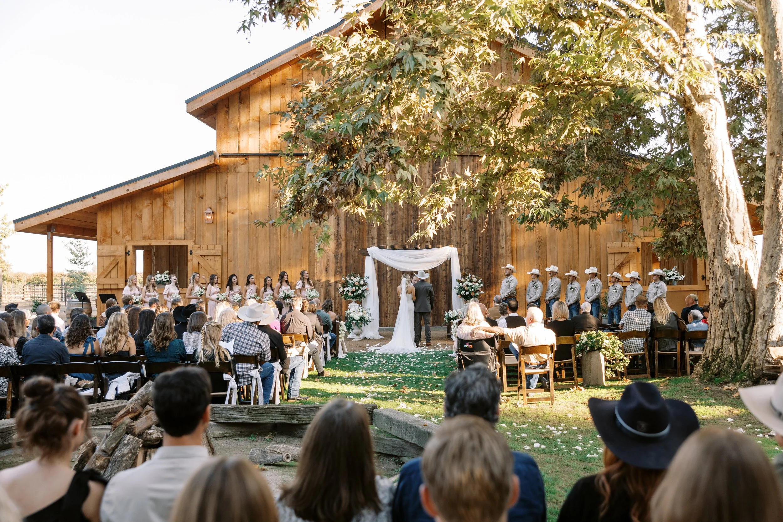 Outdoor wedding ceremony set against a rustic wooden barn, with guests seated on both sides, a bride and groom in the center, bridesmaids on the left, and groomsmen on the right, under a canopy with floral arrangements, during daylight.