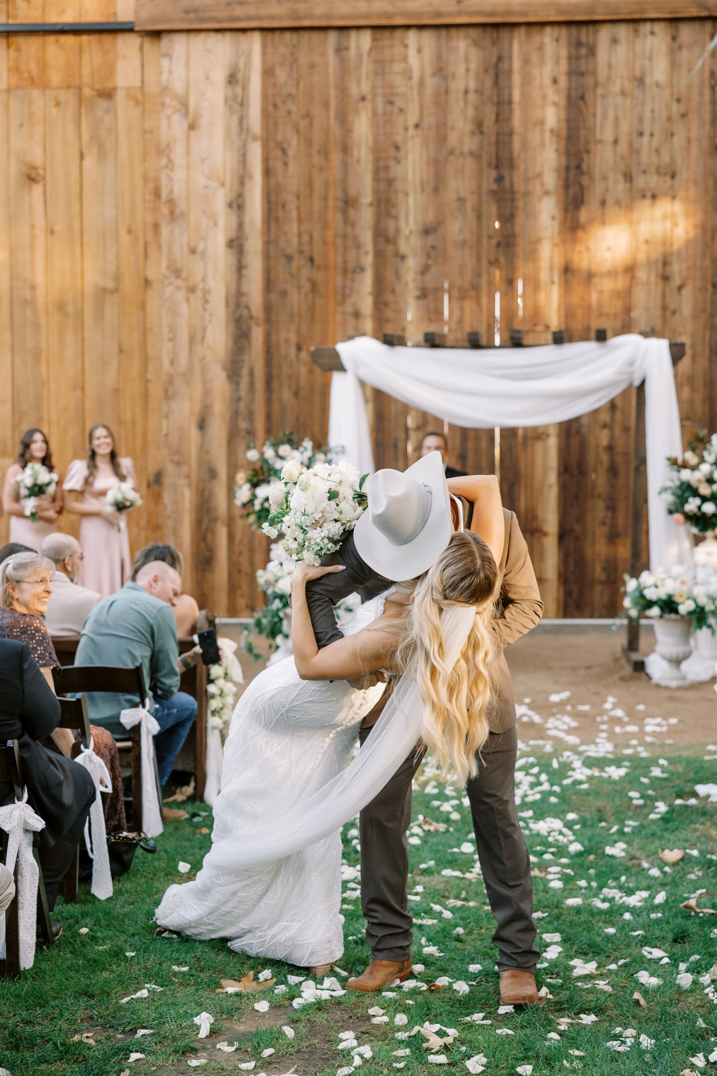 A couple is kissing during a wedding ceremony, with guests and bridesmaids in the background. The woman is wearing a white wedding dress and holding a bouquet, while the man is dressed in a cowboy hat, brown jacket, and brown boots. They are standing