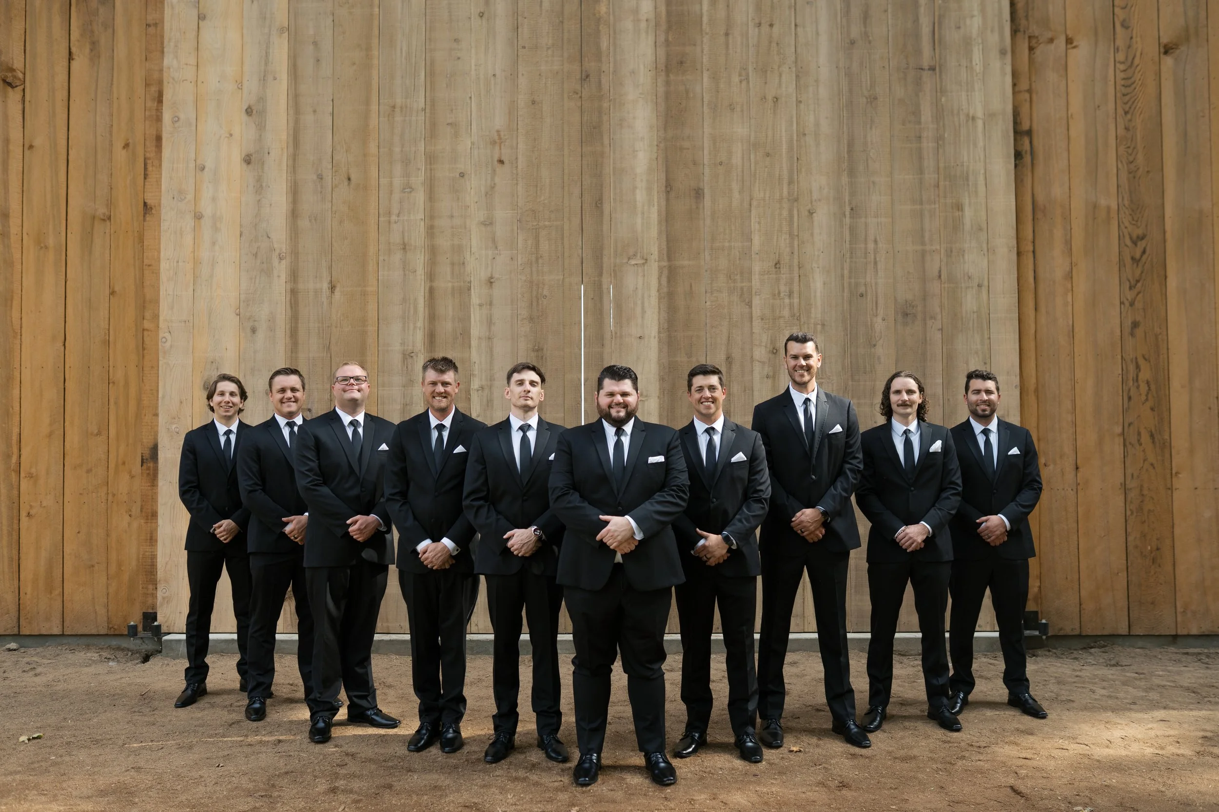 A group of ten men in formal black suits and ties standing outdoors in front of a large wooden wall.