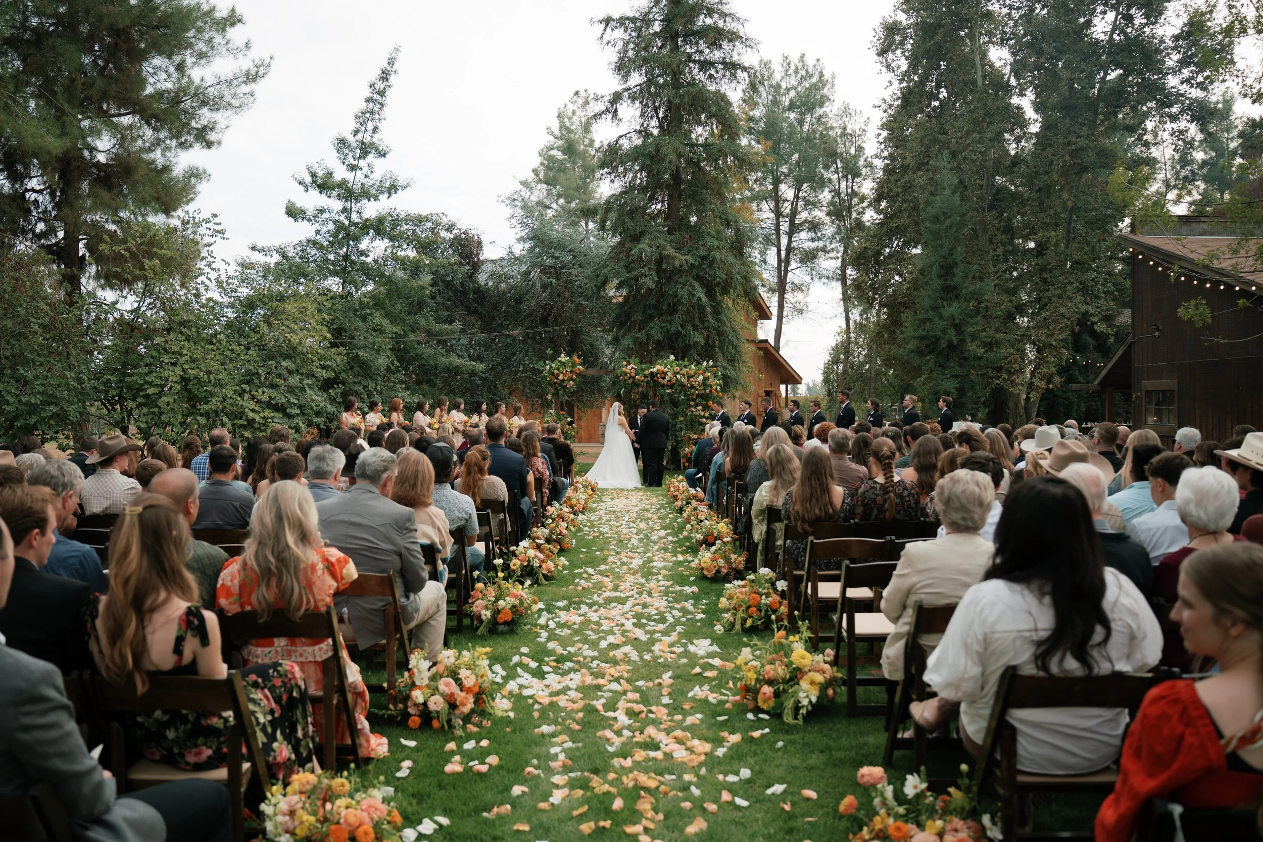 Outdoor wedding ceremony with a bride and groom standing at altar, surrounded by guests seated on both sides, trees, and floral decorations.