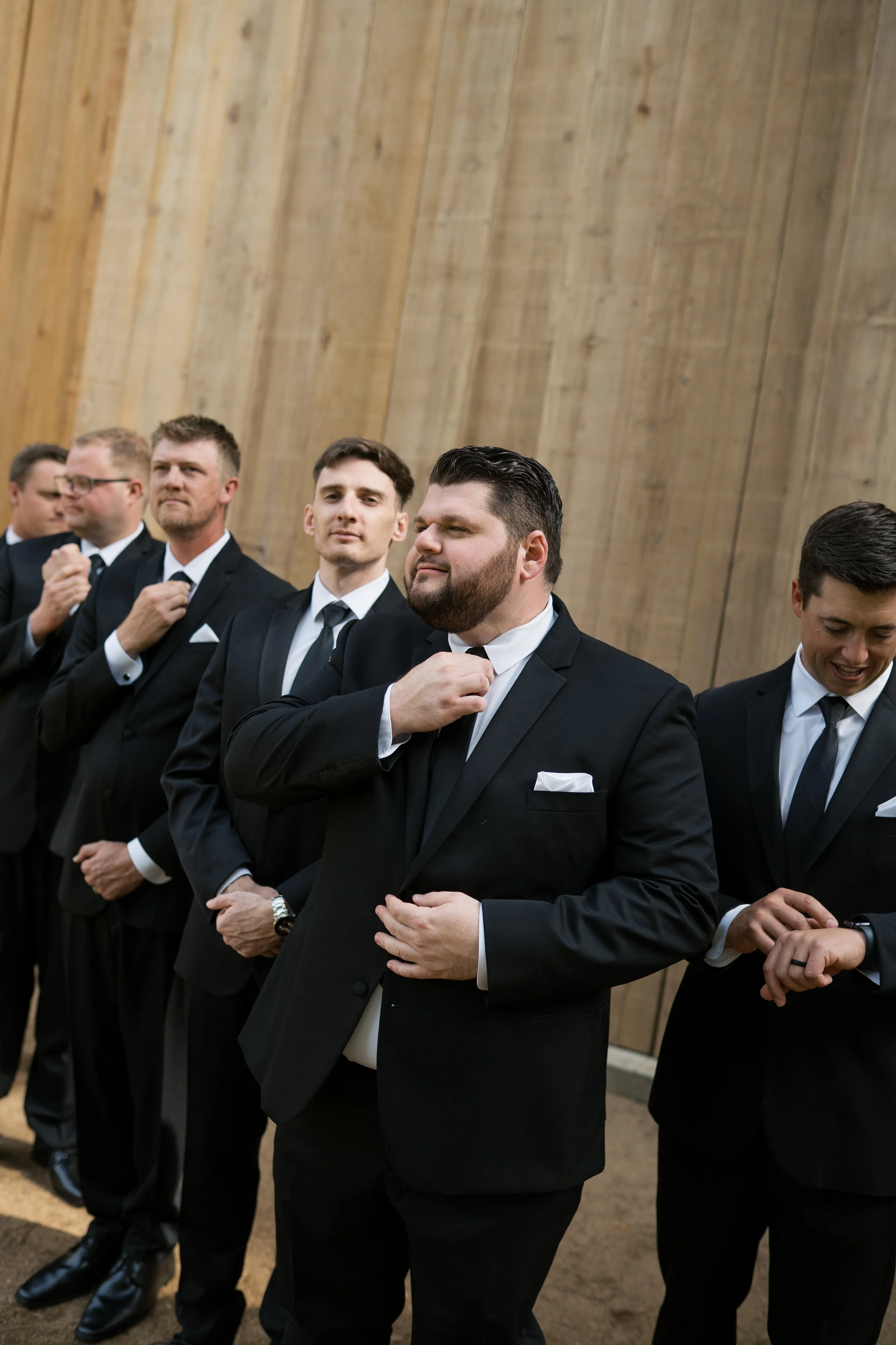 Line of men in black tuxedos adjusting their ties in front of a wooden wall, preparing for a formal event or wedding.