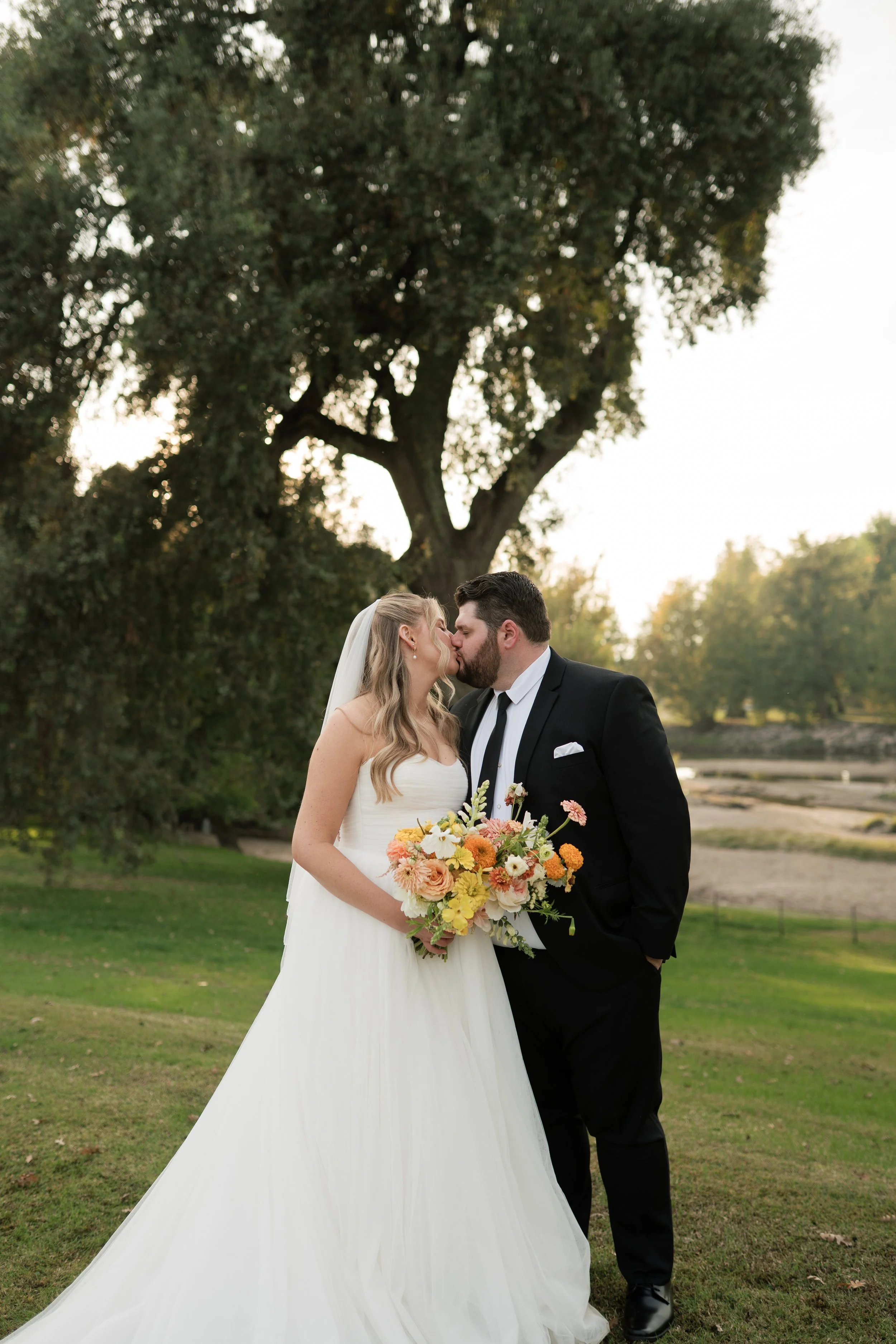 A bride and groom kiss outdoors on a grassy area with trees in the background. The bride wears a white wedding dress and veil, holding a bouquet of colorful flowers. The groom is dressed in a black suit with a white shirt and black tie.