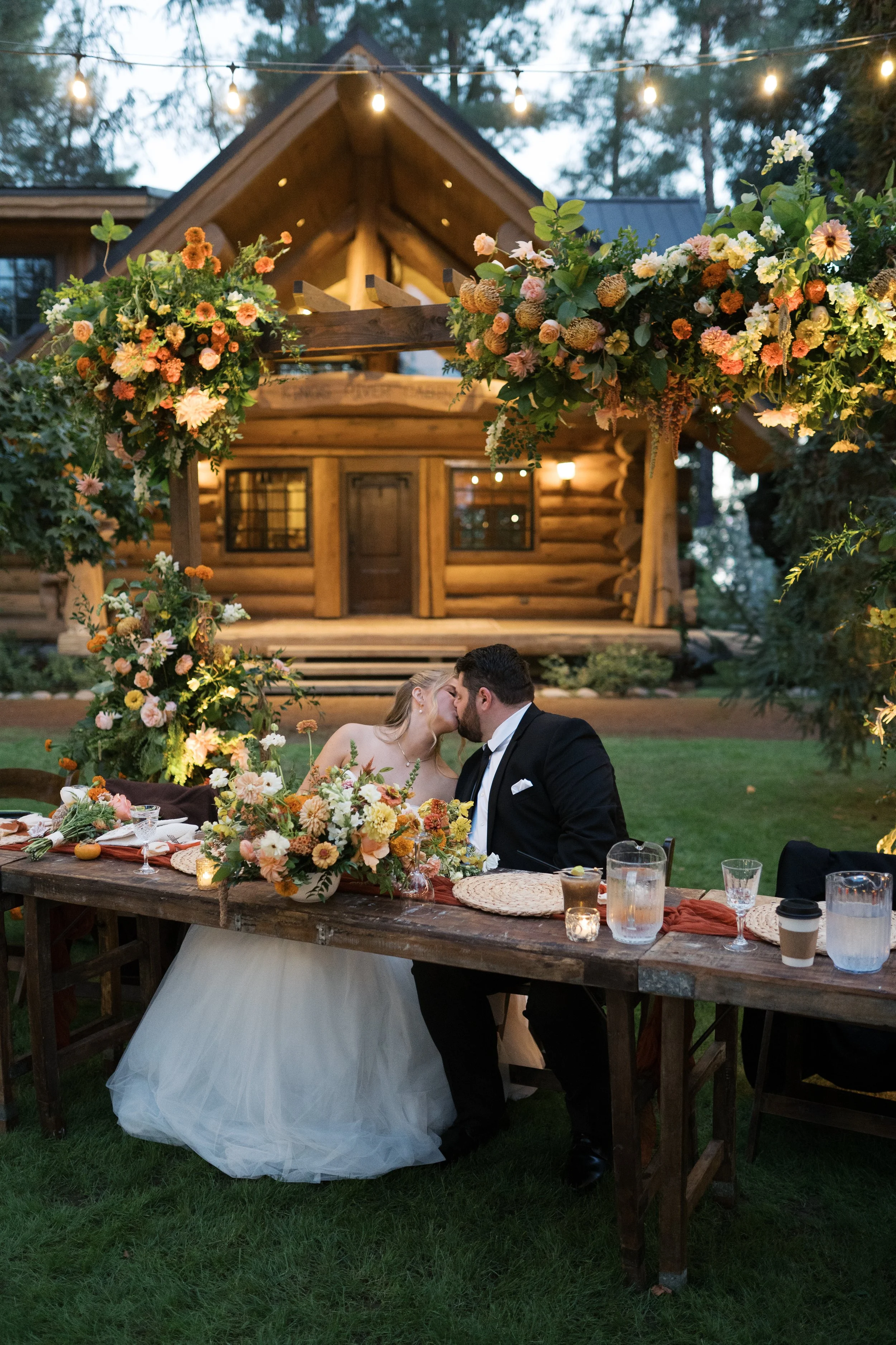 A newlywed couple sharing a kiss at a decorated outdoor wedding reception table in front of a wooden cabin, with string lights overhead and flower arrangements on the table and arch.