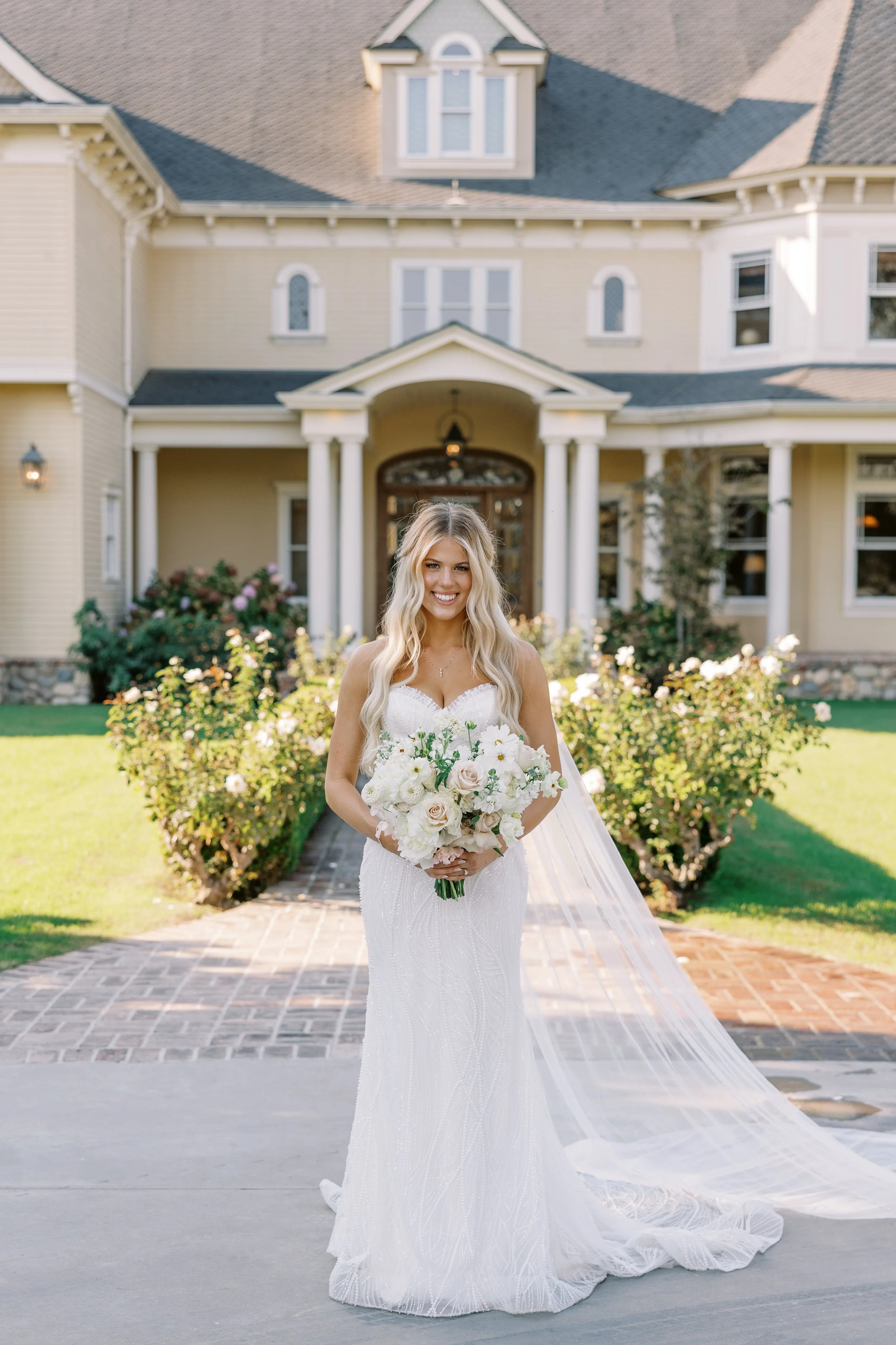 Brunette bride in a white wedding dress holding a bouquet of white and blush flowers, standing in front of a large house with a garden.